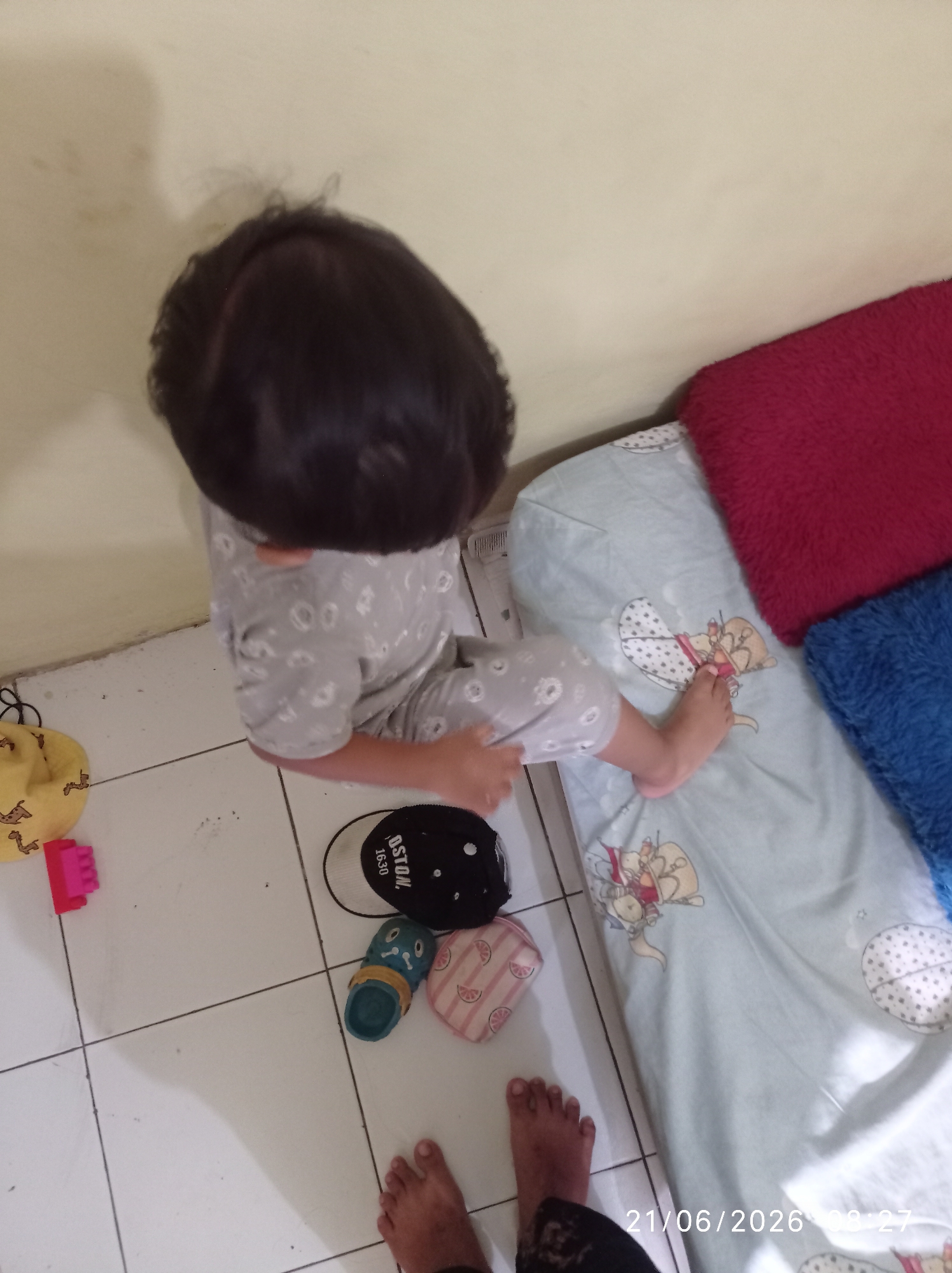 An overhead, eye-level shot shows a young child, wearing a grey and white patterned pajama set, sitting on a tiled floor. The child's dark hair is short and obscures their face. To the child's right, a bed is visible, covered with a light blue sheet featuring cartoonish drawings of animals and flowers. Two plush pillows, one red and one blue, are placed on the bed. Scattered on the floor around the child are several items: a yellow beanbag with giraffe patterns, a pink toy block, a black baseball cap with white lettering, a green and blue sandal, and a small pink and white patterned pouch. The background consists of a cream-colored wall. The lighting suggests an indoor setting, possibly a bedroom. The timestamp "21/06/2026 00:27" is visible in the lower right corner. The scene appears to be a domestic environment in the city of Sukaraja, Indonesia, with the child engaged in play or resting.