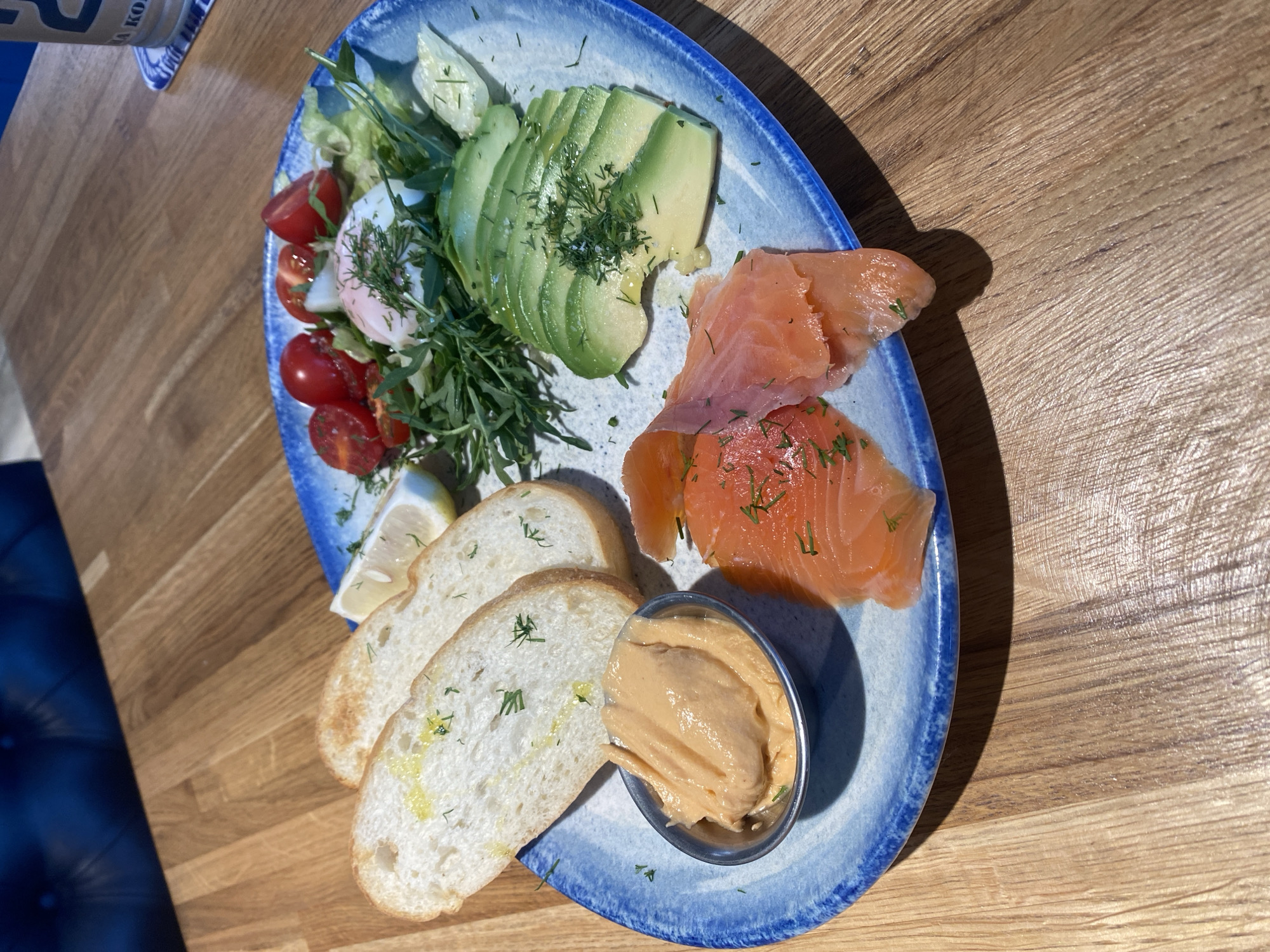 The image shows a close-up view of a breakfast platter on a wooden table. The platter, with a blue rim, contains several food items: three slices of bread topped with oil and herbs, a small metal dish filled with a creamy, orange-colored spread, slices of smoked salmon, sliced avocado, a poached egg on a bed of greens, halved cherry tomatoes, and a lemon wedge. Fresh dill is sprinkled over several of the food items. In the background, a portion of a dark blue upholstered seat and a wooden surface are visible. The setting appears to be a restaurant or cafe. The image is taken from a slightly elevated angle. There are no people visible in the image. The location is identified as Красная Пахра, Russia.