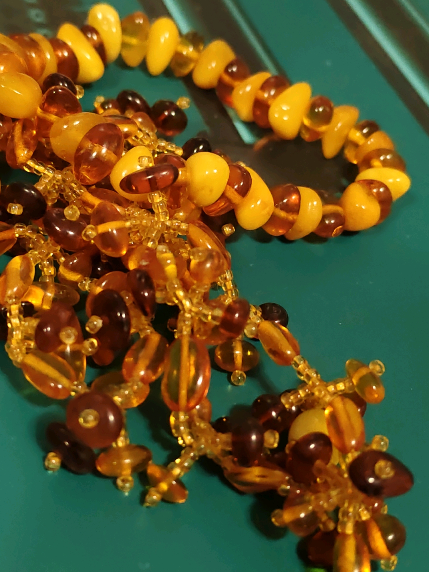 A close-up, overhead shot captures a strand of amber beads against a teal surface. The beads vary in size, shape, and color, ranging from a pale yellow to a deep, reddish-brown, with many exhibiting translucent qualities and internal inclusions. Some beads are smooth and oval-shaped, while others are more irregular or faceted. The strand is loosely coiled and draped, with a cluster of smaller, clear amber-colored beads forming a fringe at the bottom. The teal surface appears to have faint parallel lines, possibly indicating a textured material or background. There is no indication of Rostov-on-Don, Russia, or any other specific location in the image.