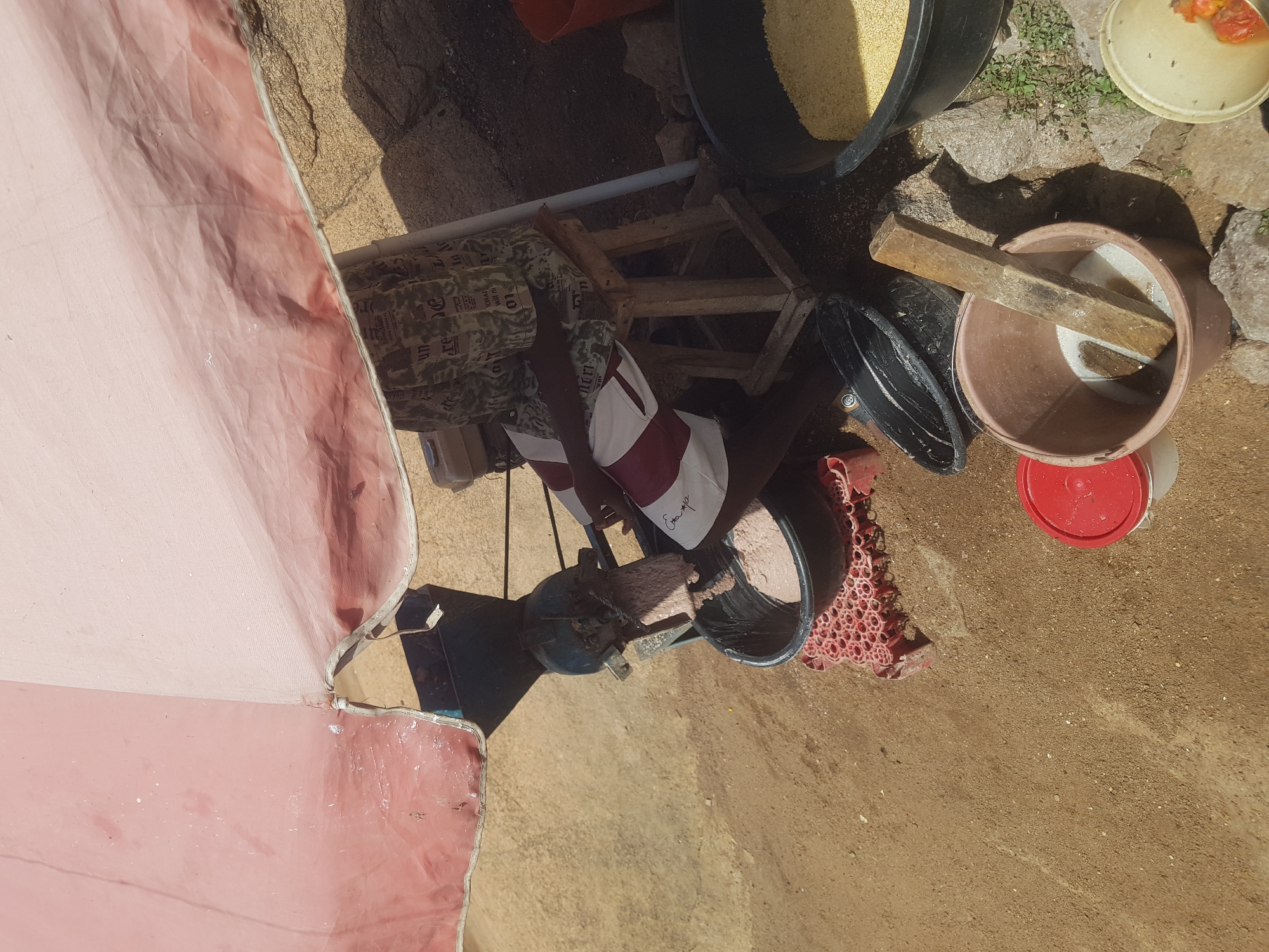 An overhead, slightly tilted, outdoor shot shows a person sitting on a wooden stool under a large, pink umbrella. The person is wearing a camouflage patterned shirt and a white shirt with red stripes. They appear to be operating a machine that grinds a light pink substance into a large black basin. Surrounding the basin is a red mesh mat. To the right of the basin is another large black basin filled with yellow grain. Next to that, on the ground, is a brown bucket containing a wooden pestle and a white liquid. Further to the right, a partially visible white bowl contains some red items, possibly tomatoes, and appears to have flies. The ground is sandy and uneven, with some scattered rocks and small green plants.