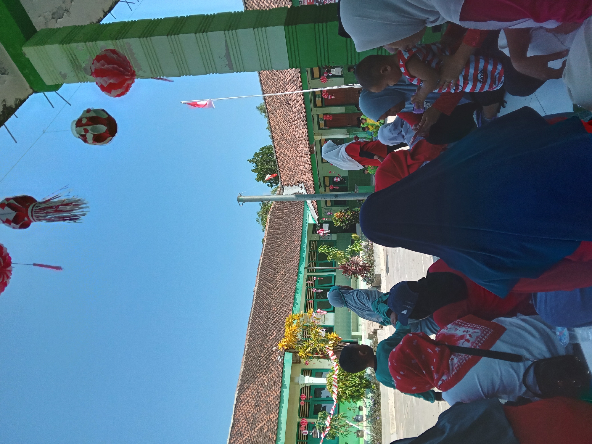 In Kedunggudel, Indonesia, on a sunny day, a group of people, mostly women and children, are gathered outdoors, likely for an event at a school. The building in the background appears to be a school with green trim and a tiled roof. Red and white decorations, including lanterns and banners, are visible, suggesting a festive occasion, possibly related to Indonesian Independence Day given the red and white colors and the Indonesian flag flying. Some individuals are seated on the ground, while others are standing. A young child in a striped shirt is being held by an adult. The overall atmosphere seems relaxed and communal.