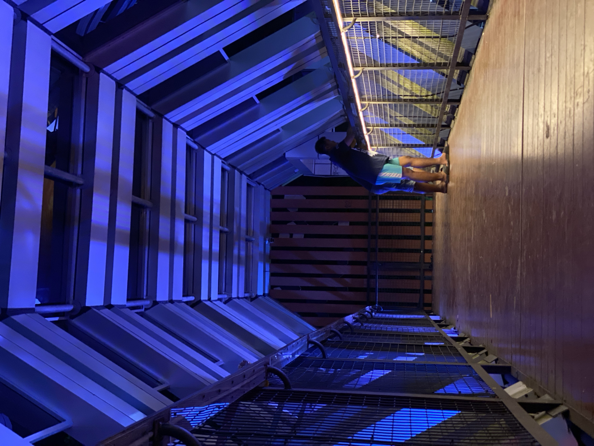 In Banjarbaru, Indonesia, two individuals are standing on a wooden elevated walkway. The walkway is enclosed by metal fencing with mesh panels and is illuminated by blue lights, creating a vibrant atmosphere. Above, a geometric ceiling structure is also illuminated with blue light, casting long shadows. The walkway leads into a dimly lit area with vertical wooden slats. The individuals are dressed casually in shorts and t-shirts, with one leaning on the railing, appearing to observe the surroundings. The overall scene suggests an outdoor recreational or architectural feature within the city.