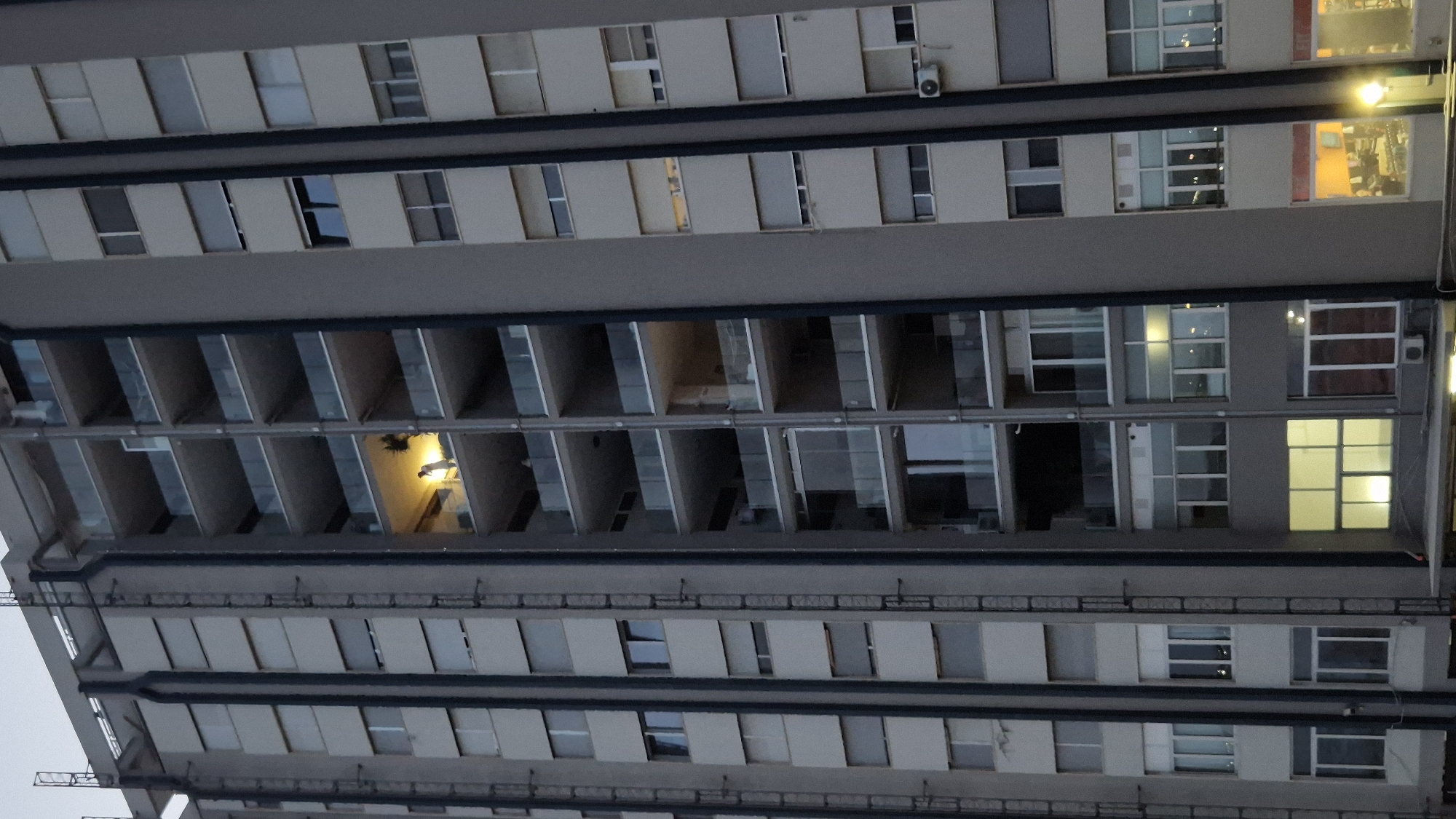 An exterior vertical shot shows a tall, multi-story apartment building in Taranto, Italy. The building has a modern design with many balconies. On one of the balconies, a person is standing and illuminated by a light source, appearing to be indoors. The lighting indicates it is dusk or night.