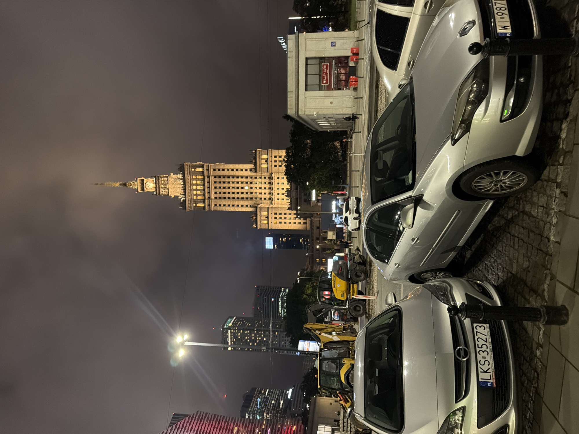 In an urban night scene, a tall, illuminated skyscraper with a clock tower stands prominently against a cloudy sky. In the foreground, several silver cars are parked on a cobblestone street. Two yellow construction vehicles, a backhoe and an excavator, are positioned behind the cars. A small, white, modern building with red signage is visible to the right of the frame. The overall impression is a city street at night, with a mix of modern architecture, construction, and parked vehicles.