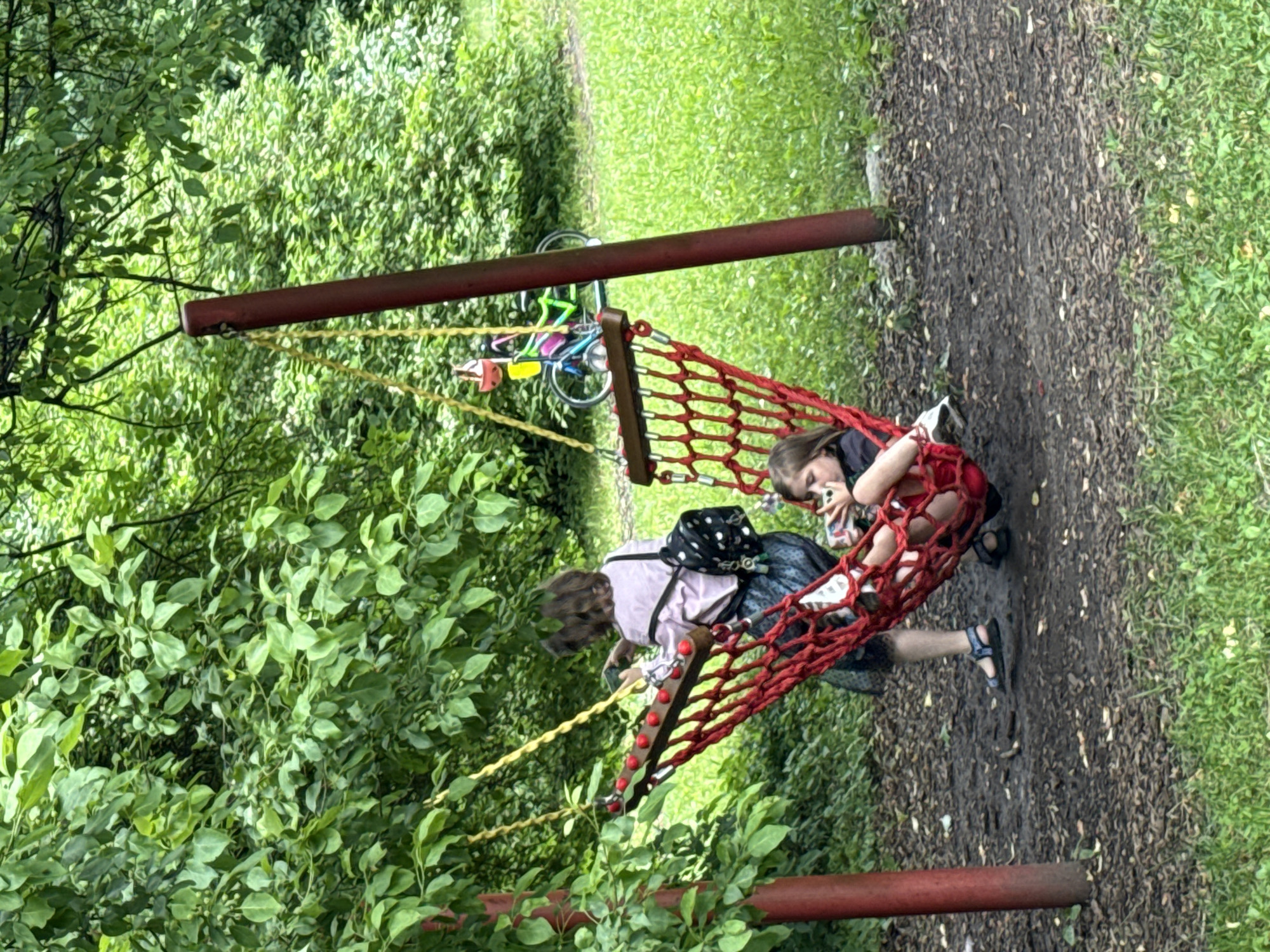 In a park setting in Moscow, Russia, two young girls are relaxing in a large red net hammock. The hammock is suspended between two red poles, which appear to be part of a playground structure. Lush green trees and foliage surround the hammock, with a dirt path and green grass visible in the background. One girl is lying down, looking at her phone, while the other is sitting up, also holding a phone. Their casual posture and the presence of the phones suggest a moment of leisure and relaxation. A bicycle with colorful accents is visible in the background, leaning against a tree, hinting at a recreational atmosphere. The lighting suggests it might be daytime, possibly overcast due to the soft light and lack of strong shadows. The overall mood is peaceful and relaxed. There is no visible text in the image.