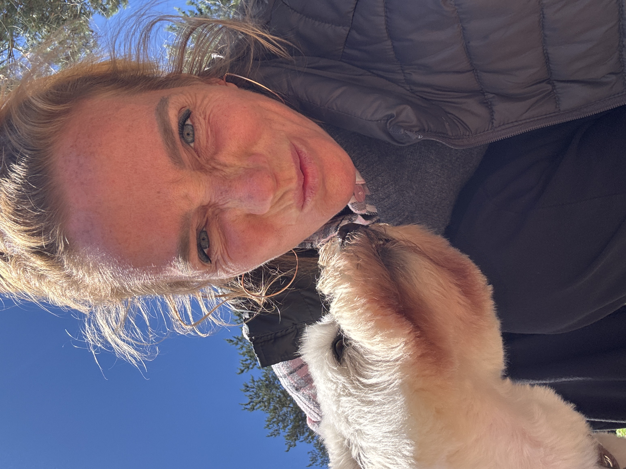 A woman with blonde hair pulled back and strands blowing in the wind takes a selfie with a white dog. She is making a silly face, scrunching up her nose and pursing her lips. The dog's snout is touching her face. She is wearing a dark puffy vest over a grey sweater and a checkered scarf. The background shows a bright blue sky and trees. The image was taken outdoors in Zelenaya Roshcha, Russia.