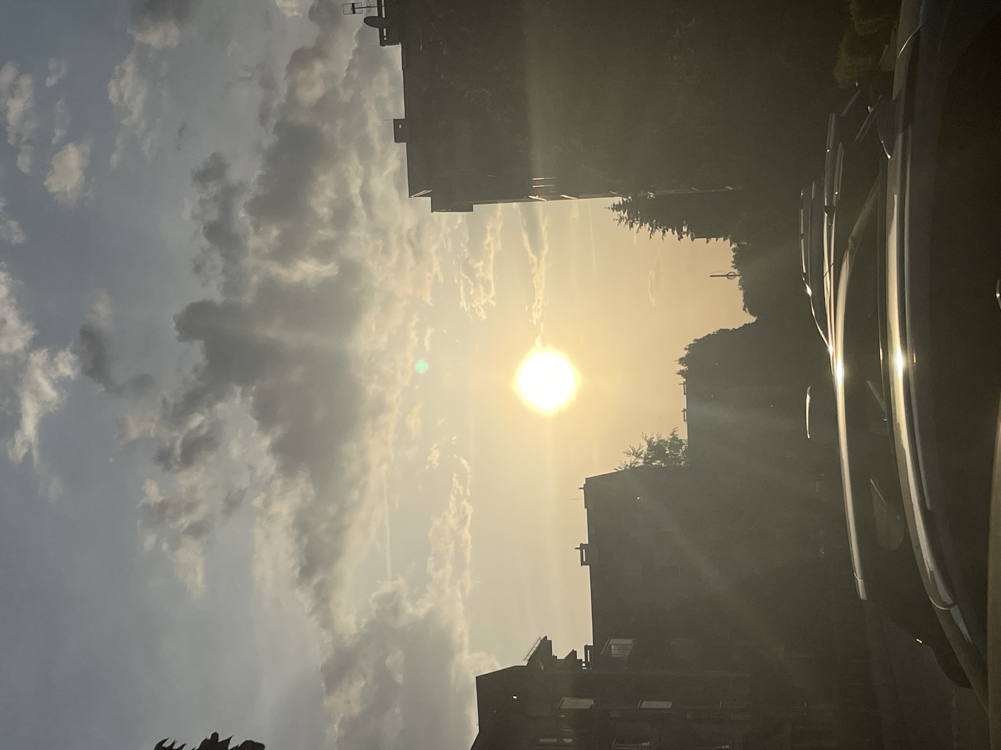The photograph depicts an outdoor scene with the sun prominently positioned in the center, surrounded by a hazy sky with light, airy clouds. Buildings flank both sides, their details obscured by the bright light and shadows. A portion of a vehicle, possibly a car, is visible on the right edge of the image, with reflective surfaces catching the light. The location appears to be an urban area due to the presence of buildings.