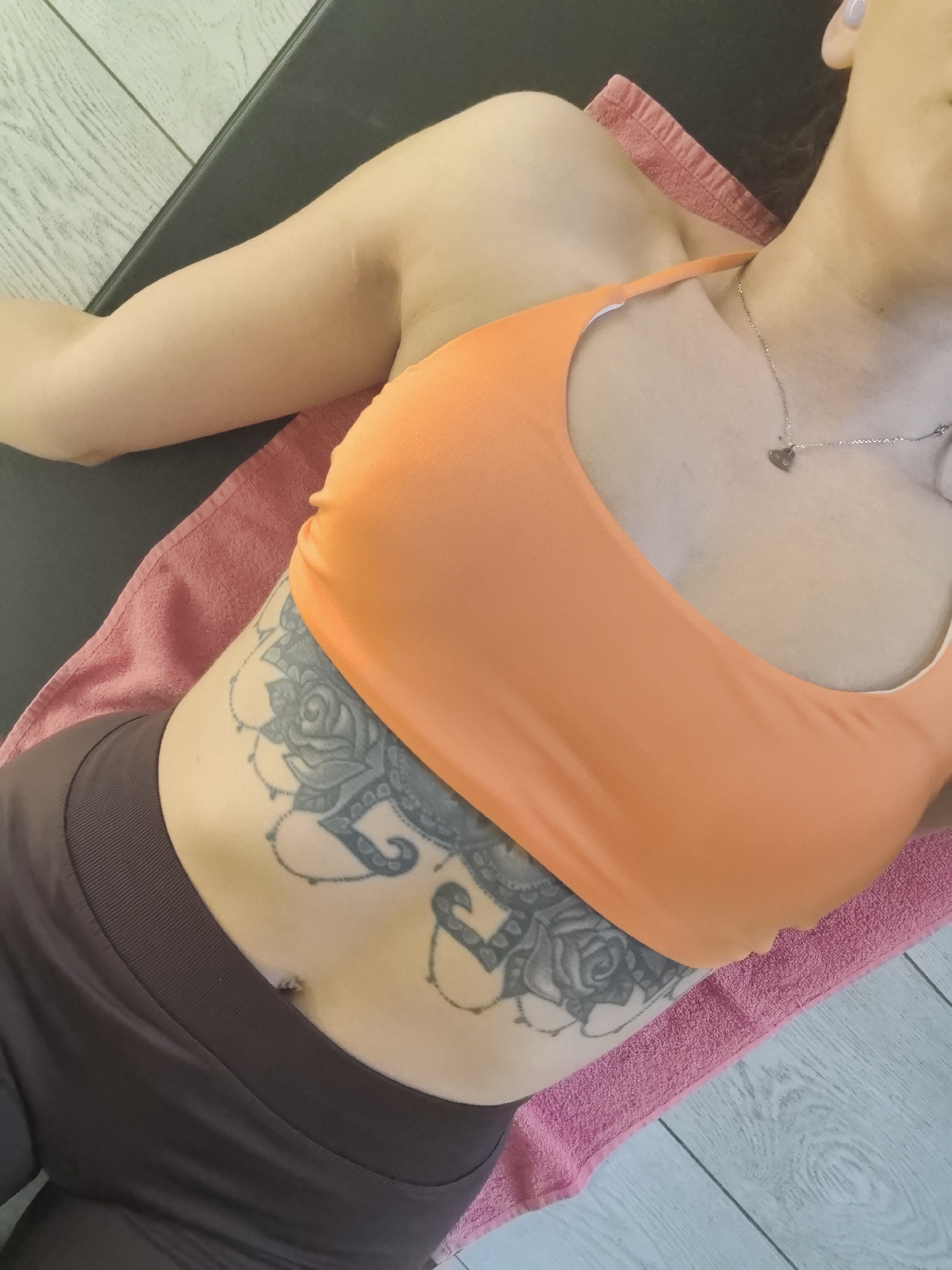 A woman lies on a pink towel, wearing an orange sports bra and brown athletic pants. A large tattoo covers her left abdomen, featuring floral and paisley patterns in gray ink. She wears a delicate silver necklace with a heart pendant. She rests on what appears to be an exercise mat, with a white wooden floor visible in the background. The location cannot be determined as Stara Zagora, Bulgaria without further information.