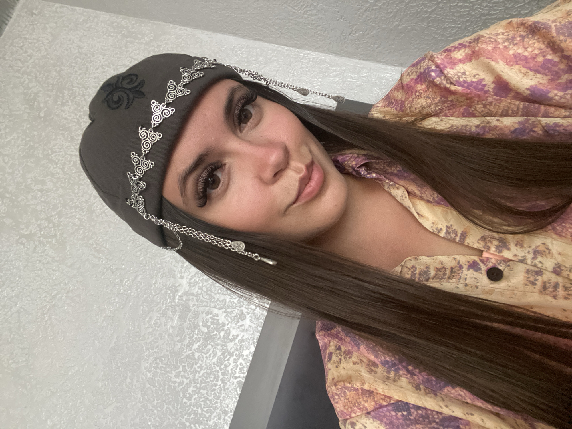 A young woman with long, dark brown hair is looking at the camera with a slight smile. She is wearing a grey beanie with decorative silver chains and charms that hang down her forehead and near her ears. She is also wearing a patterned shirt with colors of yellow, pink, and purple. The background is a textured grey wall. The image appears to be taken indoors. There is no information available about the location in the city of Chalda, Russia.