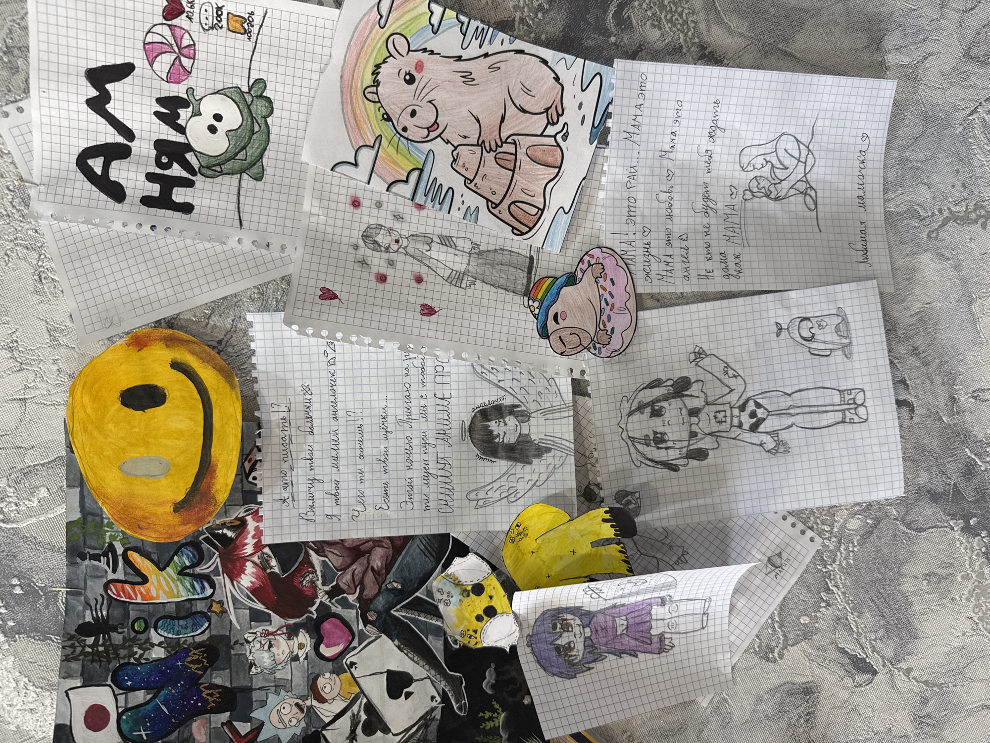 The image is an eye-level, overhead shot of a collection of drawings and notes scattered on a surface. The drawings, mostly done on graph paper, depict various cartoon-like characters and figures. Among them are a green creature, a bear standing on an object near a rainbow, an anime-style figure with wings, and other animated characters. The notes are handwritten in what appears to be the Russian language. One note expresses love for "Mama," while others contain phrases like "I am your sweet angel."
A yellow, round object resembling a smiley face is situated near the bottom-left corner of the image. Below it is a paper with anime-style stickers and a Japanese flag.
The exact location within the city of Rusanovo, Russia, cannot be determined from the image.