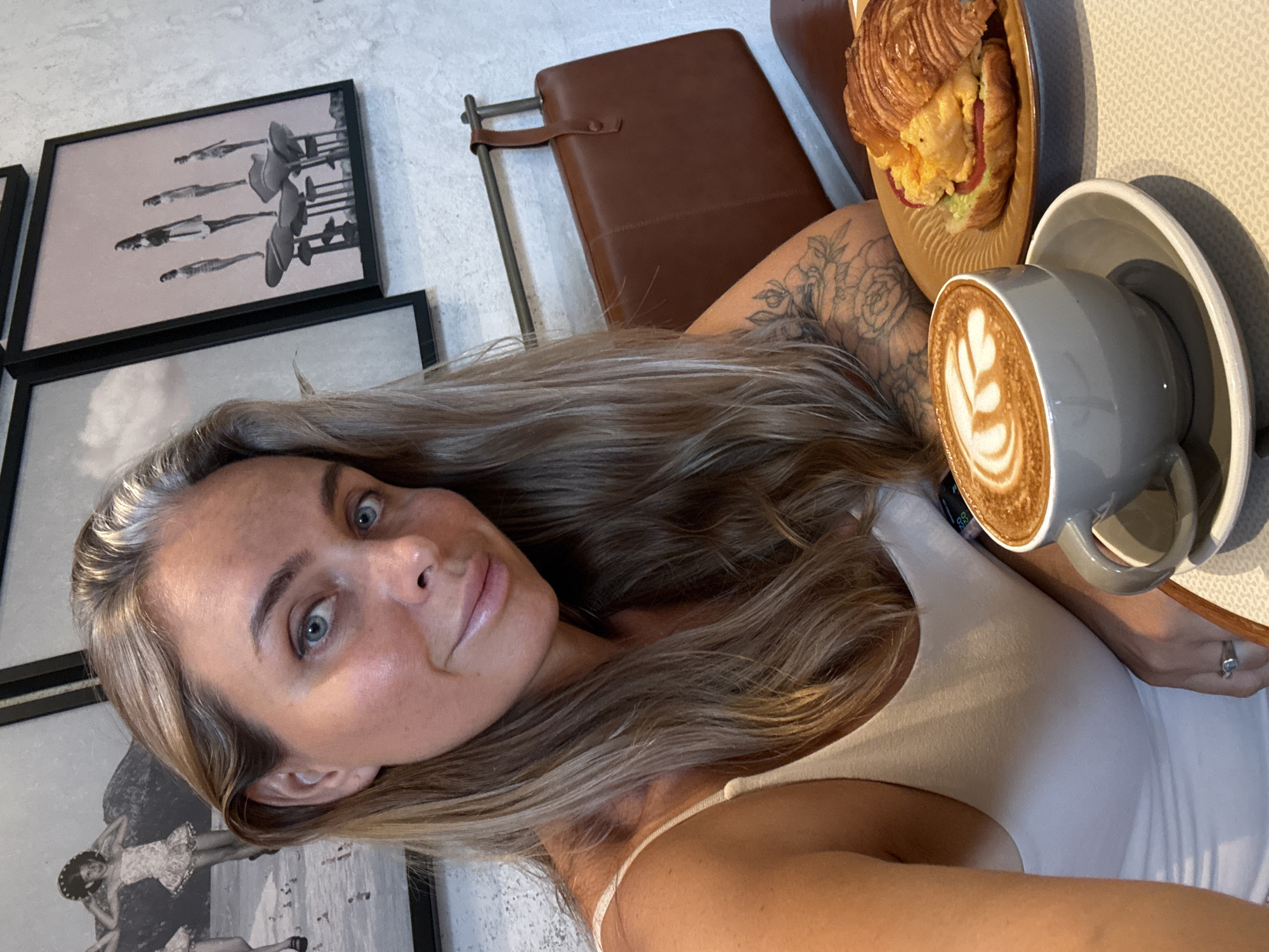 A woman with blonde hair and blue eyes is taking a selfie in a cafe. She is wearing a white tank top and has tattoos on her arm. In front of her is a cup of coffee with latte art and a croissant sandwich. The cafe has framed pictures on the wall, including one with several figures standing on mushroom-like objects. The overall atmosphere suggests a modern and trendy establishment in Shanghai, China.