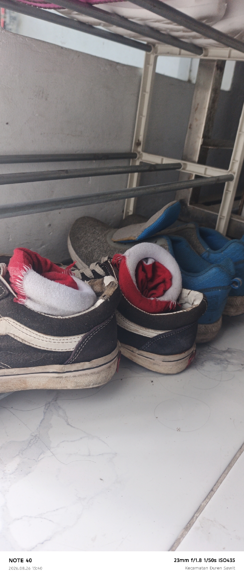 The image shows a close-up, low-angle shot of several pairs of shoes on a tiled floor. In the foreground, a pair of dark Vans high-top sneakers with white side stripes is visible. One shoe has a red and white sock stuffed inside. Next to it, another similar shoe is slightly out of focus, also with a red and white sock. Behind these, a grey wool sneaker with a blue and grey sole is partially visible. Further back, a pair of bright blue sneakers is in focus. The shoes appear to be on a drying rack or shelf unit with metal bars and white plastic supports. The background is a light-colored, possibly concrete, wall. The floor has a white marble pattern with some scattered debris and hair. The metadata indicates the image was taken with a "NOTE 40" device on "2026.08.26 13:40" with "23mm f/1.8 1/50s ISO435" and geotagged to "Kecamatan Duren Sawit," which is located in East Jakarta, Indonesia.