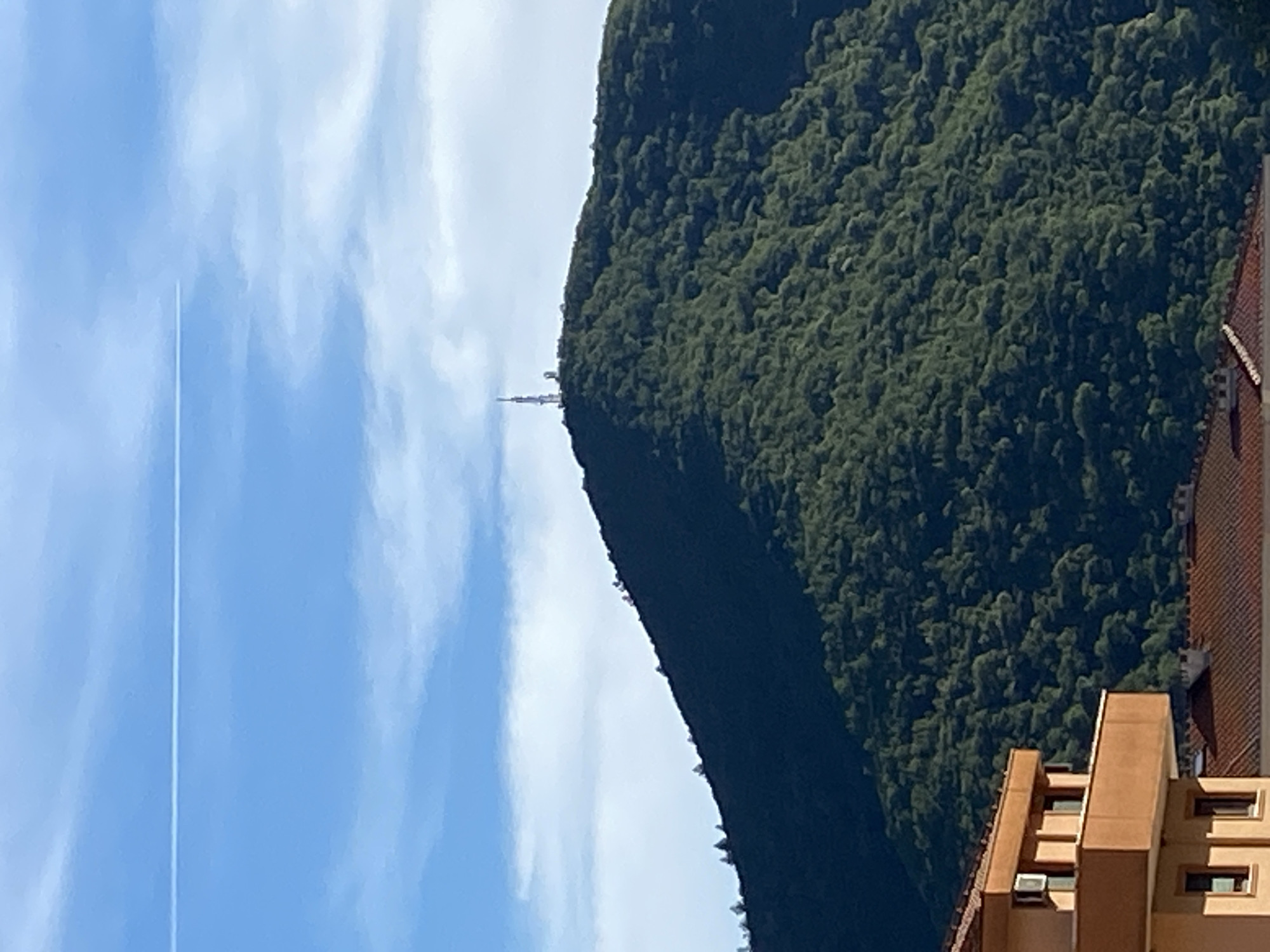 The image shows a telecommunications tower on top of a densely forested mountain. The mountain slope is covered with dark green trees, and a few scattered trees are visible on the ridge. The sky above is a clear blue with some wispy white clouds. A faint white contrail from an airplane stretches horizontally across the upper portion of the sky. In the foreground, the upper part of a brown building with a balcony and windows is visible, along with the tiled roofs of other buildings. The scene appears to be a view from Budva, Montenegro, overlooking the surrounding landscape. There are no people or specific actions depicted in the image.