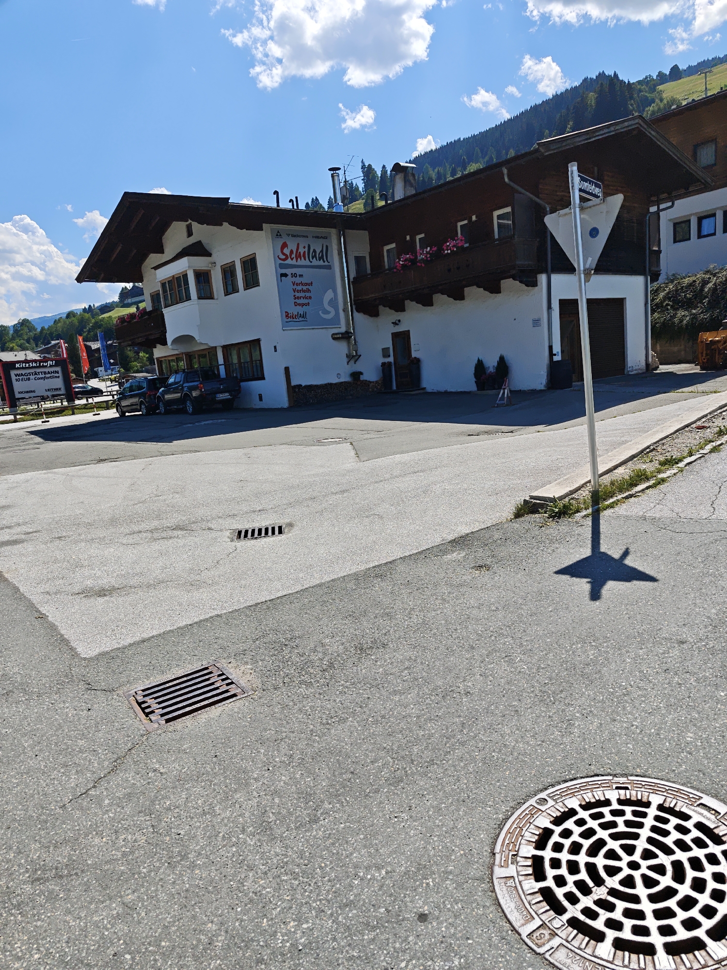 A low-angle, eye-level, exterior shot captures a sunny day in Filzen, Austria, focusing on a building with an attached advertisement for "Schiladl." The building's facade is white with dark wooden eaves and balconies adorned with pink flowers. The advertisement, a large blue and white banner, lists services such as "Verkauf," "Verleih," "Service," and "Depot" alongside the text "Schiladl."
In the foreground, a paved road with a manhole cover is visible. The road leads to the building, where two cars, a dark SUV and a black pickup truck, are parked. To the left of the building, a sign displays information about the "Wagstättbahn" cable car.
Behind the building, a tree-covered hillside rises, culminating in a clear blue sky with scattered white clouds. A triangular street sign indicating "Sonnfeldweg" is also present. The scene suggests a commercial area in a mountainous region.