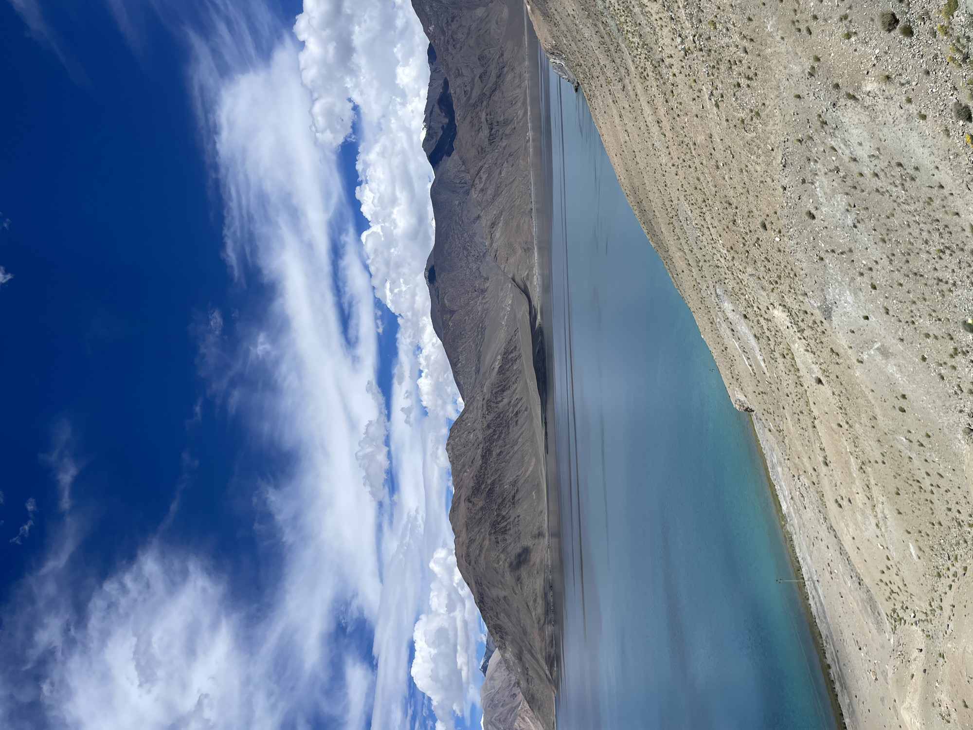 The image depicts a wide-angle view of a serene lake nestled amidst rugged, barren mountains under a brilliant blue sky dotted with fluffy white clouds. The lake's water exhibits a striking gradient of blue hues, transitioning from a deeper, more opaque tone in the distance to a translucent turquoise near the shore. The mountainous terrain is characterized by dry, rocky slopes, with sparse vegetation clinging to the earth. The foreground features a steep, sandy incline, sparsely covered with small bushes and rocks, leading down to the lake. The sky dominates the upper portion of the image, with the sun casting subtle shadows on the mountain peaks. The scene is devoid of any human presence or man-made structures, emphasizing the raw, natural beauty of the landscape. It is a clear day with good visibility, suggesting a dry and arid climate typical of high-altitude regions. The overall impression is one of vastness and tranquility.