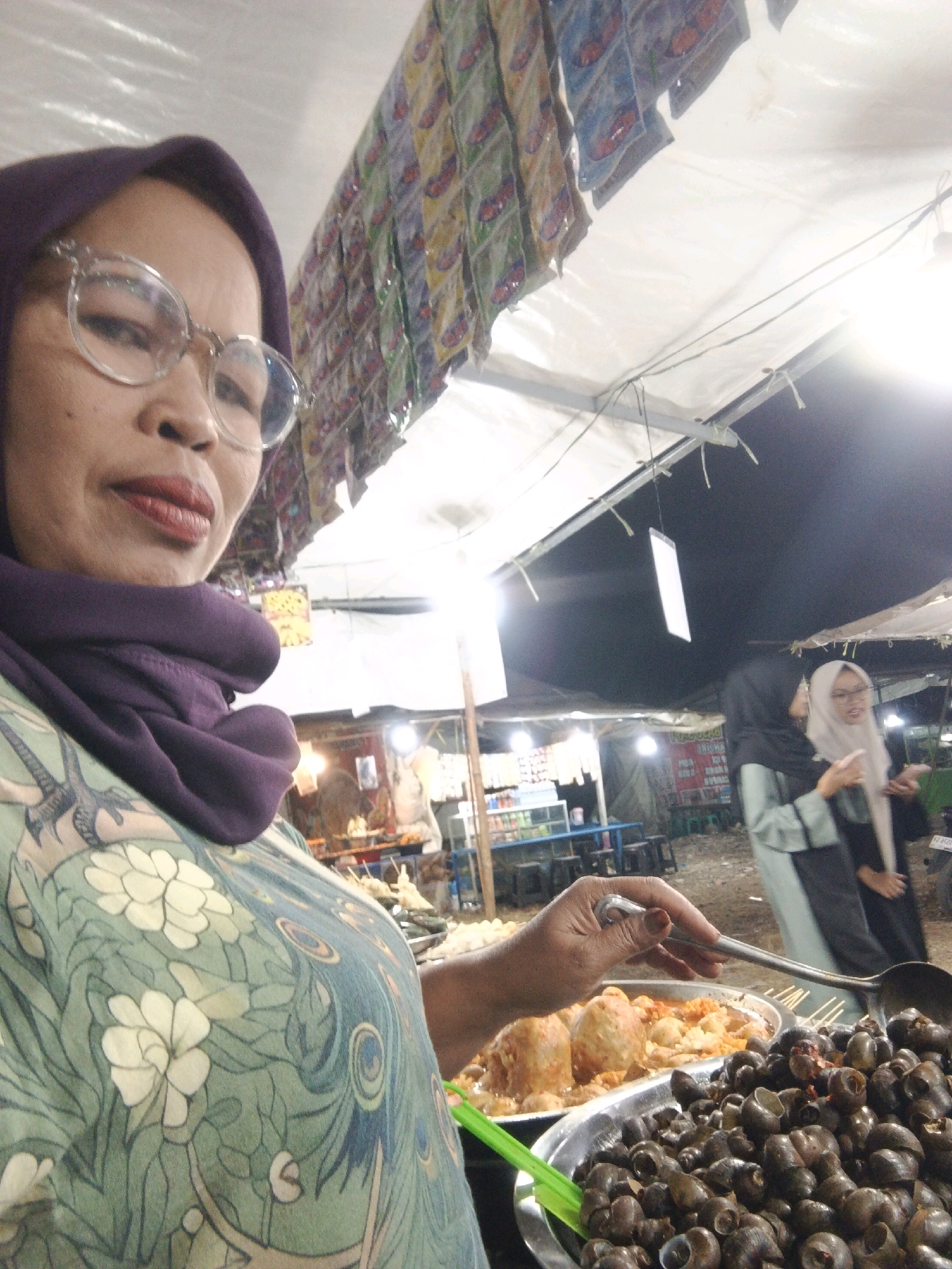 The image shows a close-up of a woman wearing glasses and a purple hijab, looking into the camera. She is standing at an outdoor food stall, with various food items displayed in front of her. In the foreground, there are two large bowls of food. The bowl on the right is filled with small, dark-shelled snails, and a ladle is placed on top. The bowl on the left contains larger, round food items, possibly meatballs or dumplings, in a sauce. Behind the woman, other food stalls and people are visible, suggesting a bustling market or street food area. The overall setting appears to be at night or in low light conditions, with artificial lighting illuminating the scene. The woman is wearing a light green and cream floral patterned shirt. The backdrop includes a white canopy, strings of what appear to be packaged snacks or goods, and blurred figures of other individuals. The environment suggests a typical street food scene in a city like Sukabumi, Indonesia.