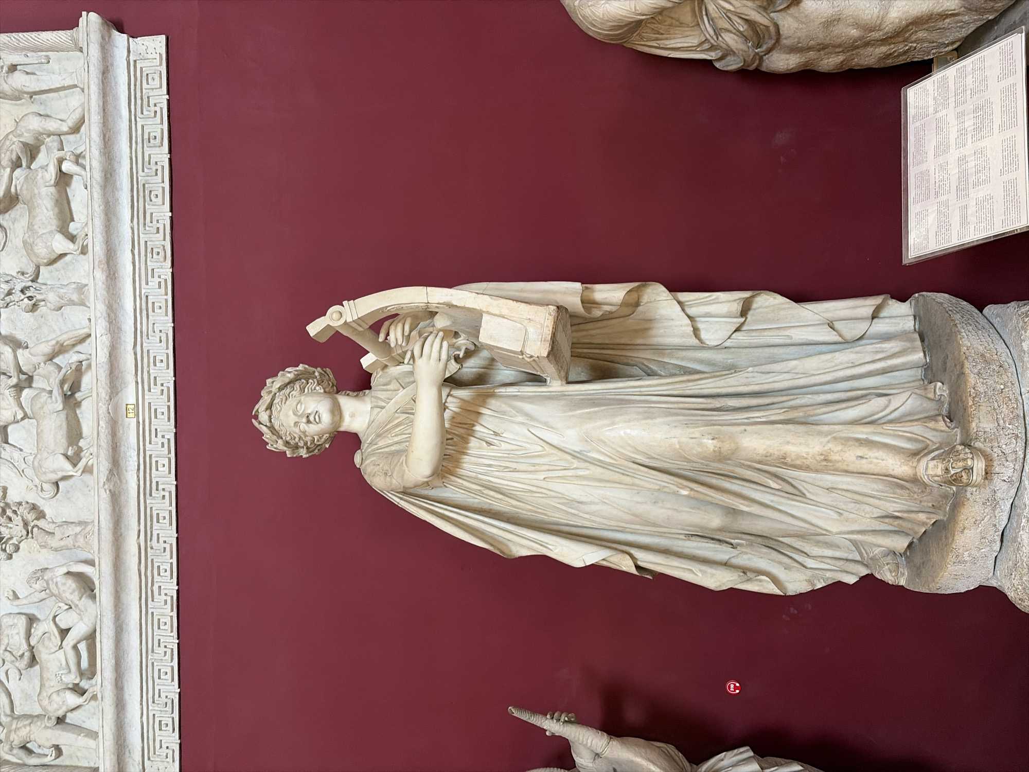 The image shows a marble statue of a woman holding a lyre against a deep red wall. The statue is a full-body, standing depiction of a female figure wearing a draped garment and a laurel wreath on her head. Her arms are bent, and her hands are positioned as if playing the lyre, which rests on her left arm. The statue stands on a circular stone base.
In the background, above the statue, is a relief carving of figures and animals, possibly depicting a mythological scene. Below the relief, a decorative frieze runs horizontally. To the far left of the statue, a portion of another marble sculpture is visible, and to the far right, the shoulder and head of another statue can be seen. In the lower right corner, a plaque with text is placed on the floor. The overall setting appears to be a museum or art gallery. This statue is the Aphrodite of Praxiteles, located at the Capitoline Museums in Rome, Italy.