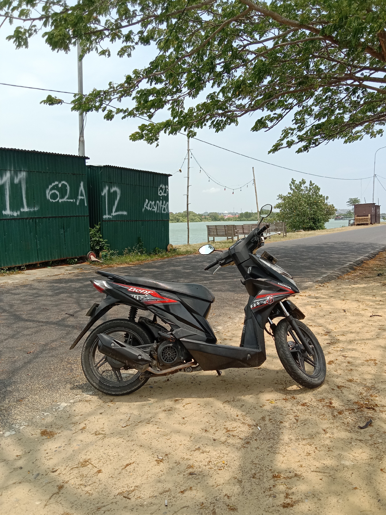 A black Honda Beat scooter is parked on a road with a sandy shoulder near the Arun Lido reservoir in Lhokseumawe, Indonesia. Behind the scooter are two green corrugated metal structures with white graffiti, reading "11 62A 12" and "62A ROSNITEK." The reservoir is visible in the background, with benches and a small structure on the opposite shore. A paved road runs alongside the reservoir, bordered by trees. Power lines and poles are present in the background. The weather appears sunny.