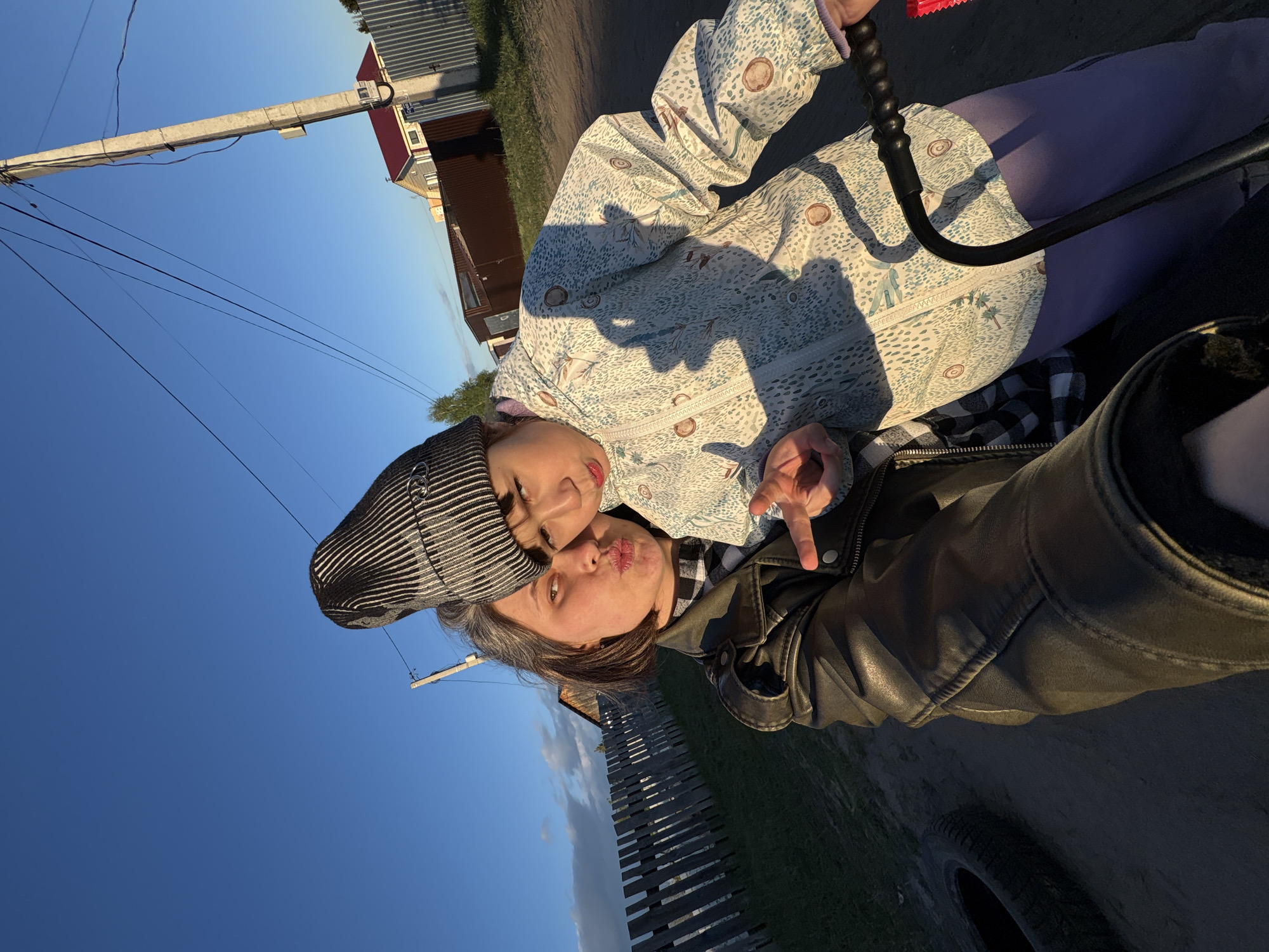 A woman and a child pose for a selfie outside in Chaikovsky, Russia. The woman, wearing a black leather jacket and a black and white striped hat, is turned slightly to the right, puckering her lips as if giving a kiss. The child, wearing a white jacket patterned with blue and brown shapes and lilac-colored pants, leans on her. The child is also puckering their lips for a kiss, and is holding up two fingers. The sky above is a clear blue, and the sun appears to be setting. A wooden fence and a few houses are visible in the background, along with a telephone pole and some wires. A tire can be seen in the foreground.