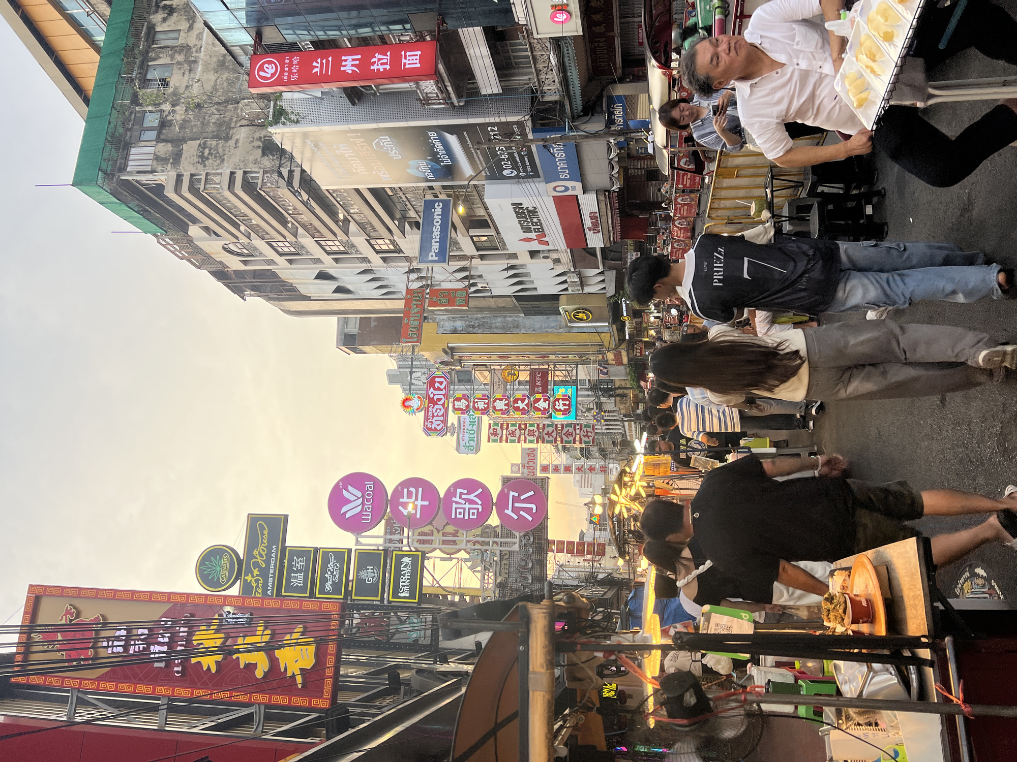 The image depicts a busy street scene in Bangkok, Thailand, likely in the Chinatown area, given the prevalence of Chinese characters on signs and the general atmosphere. The photo is taken from a low angle, looking down a street lined with multi-story buildings. Many of these buildings feature various signs in Thai and Chinese, indicating businesses such as restaurants, shops, and perhaps residences.
Several people are visible, walking in various directions, suggesting a bustling pedestrian environment. Some individuals are dressed casually, while others appear to be vendors or shopkeepers. One man in the foreground, wearing a white shirt, is seated and looking to his left, with trays of what appear to be fruit in front of him. Behind him, a group of people are walking away from the camera.
Street vendors are also present, with stalls set up along the side of the street selling food or other goods. The lighting suggests it is either early morning or late afternoon, with the sun casting a warm glow. The overall impression is one of vibrant urban life with a strong cultural influence from Chinese heritage, characteristic of Bangkok's Chinatown.