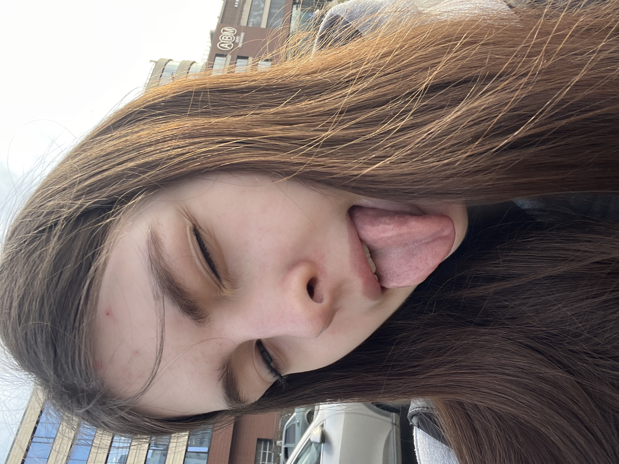 A young woman with long brown hair is seen from a low angle, sticking her tongue out and closing her eyes. In the background, there is a building with the abbreviation "ABM" visible on its facade. Based on the architecture and the "ABM" abbreviation, the location is identified as Saint Petersburg, Russia.