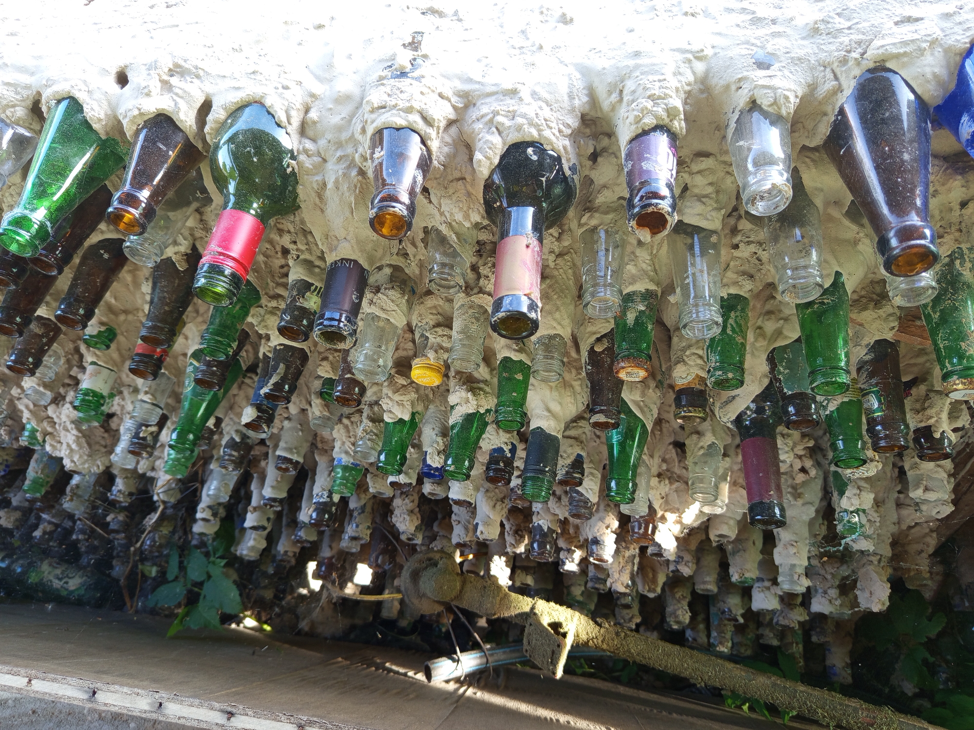 This image presents a unique architectural or artistic installation, viewed from below and looking upwards. Numerous glass bottles of various shapes, sizes, and colors (green, brown, clear, blue, and purple) are embedded into a textured, white, concrete-like material, possibly foam or plaster. The bottles are mostly broken or cut at their bases, with their necks facing downwards, creating a dense, stalactite-like canopy.
The scene is brightly illuminated by strong sunlight, indicating a sunny daytime setting. Highlights glint off the glass surfaces and the uneven texture of the white material. Below the suspended bottles, glimpses of green plant foliage and a sandy or dusty ground surface are visible, confirming an outdoor environment. A long, stick-like object, possibly a piece of wood or a tool covered in green, fuzzy material, lies horizontally in the lower part of the frame.
Notable details include the rough, organic appearance of the white embedding material. Some bottles show remnants of their original labels or caps: partial text "INK" is visible on one dark bottle, a prominent red label is on a green bottle, and a yellow cap is seen on a clear bottle. The overall impression is one of creative repurposing of discarded materials to form a distinctive structure, located in Ostrovtsy, Russia.