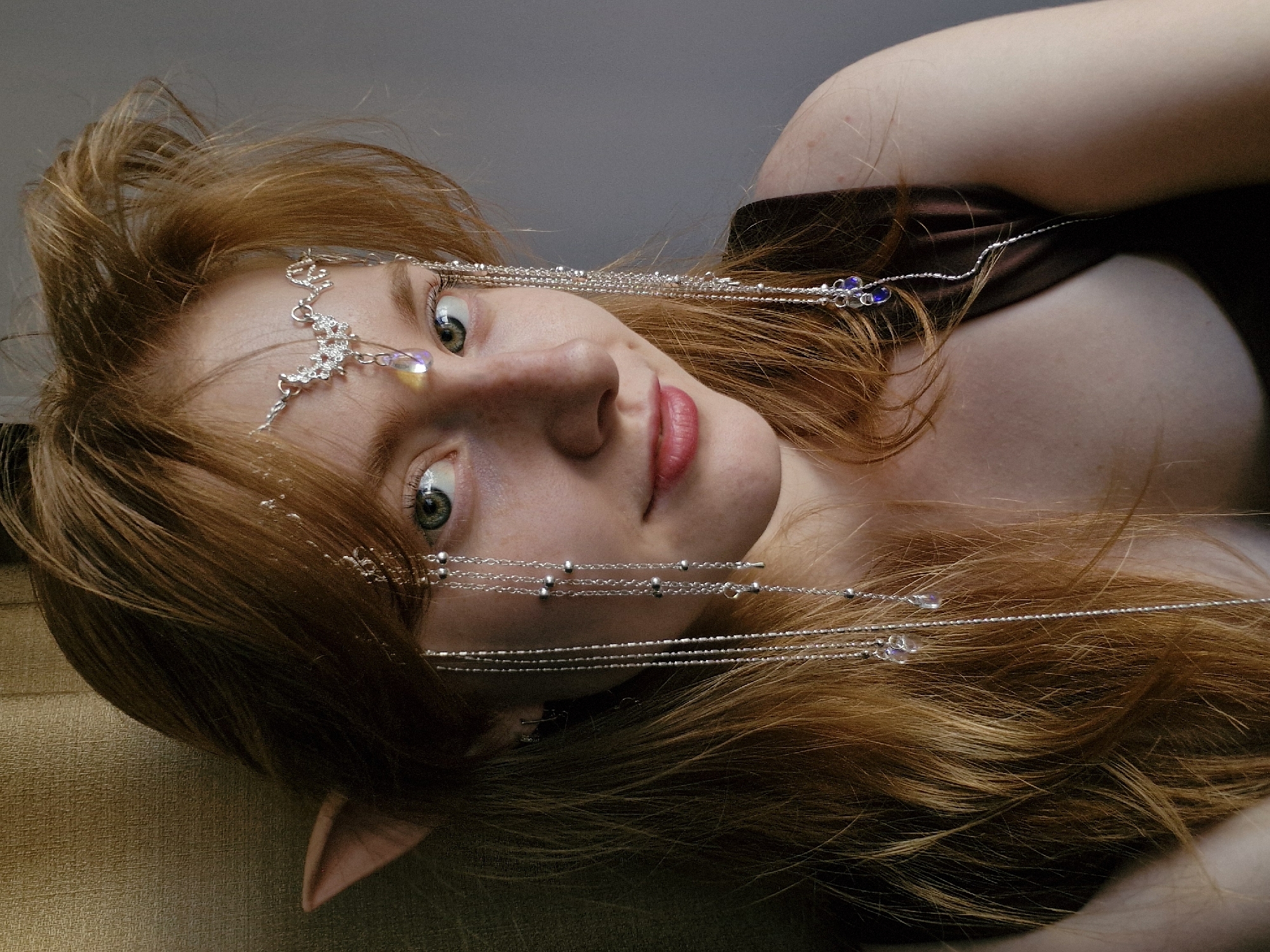 A young, fair-skinned woman with red hair is lying down, her head resting on a light brown, soft surface. She has green eyes and is wearing a silver headpiece with chains and small crystals. Her ears are pointed, suggesting an elf-like appearance. She is wearing a brown garment. The background is a muted gray. The image is taken from a low angle. There is no indication of this image being taken in Yekaterinburg, Russia.