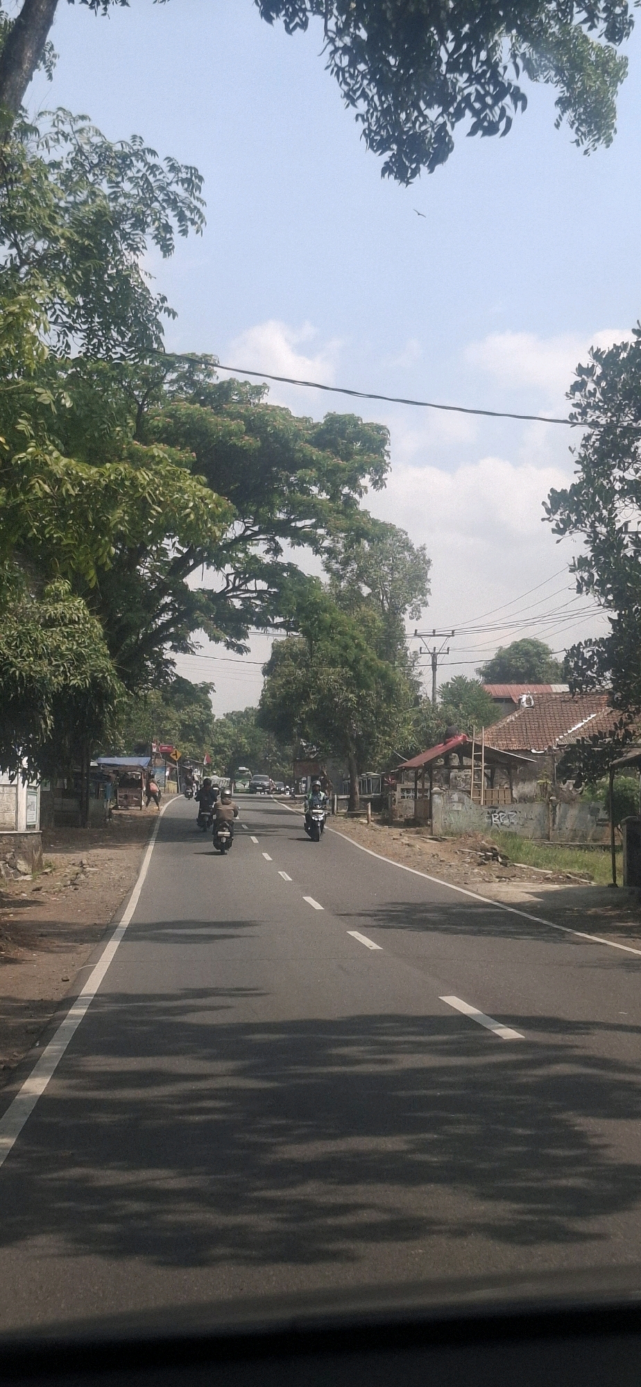 The image shows a paved road in what appears to be a rural or suburban area of Susukan, Indonesia, lined with trees and some buildings. The road has a white line marking the center and a white edge line. There are several motorcycles on the road, some with riders. In the background, there are houses with tiled roofs and what appears to be a power pole. The sky is partly cloudy. There are shadows on the road cast by the trees.