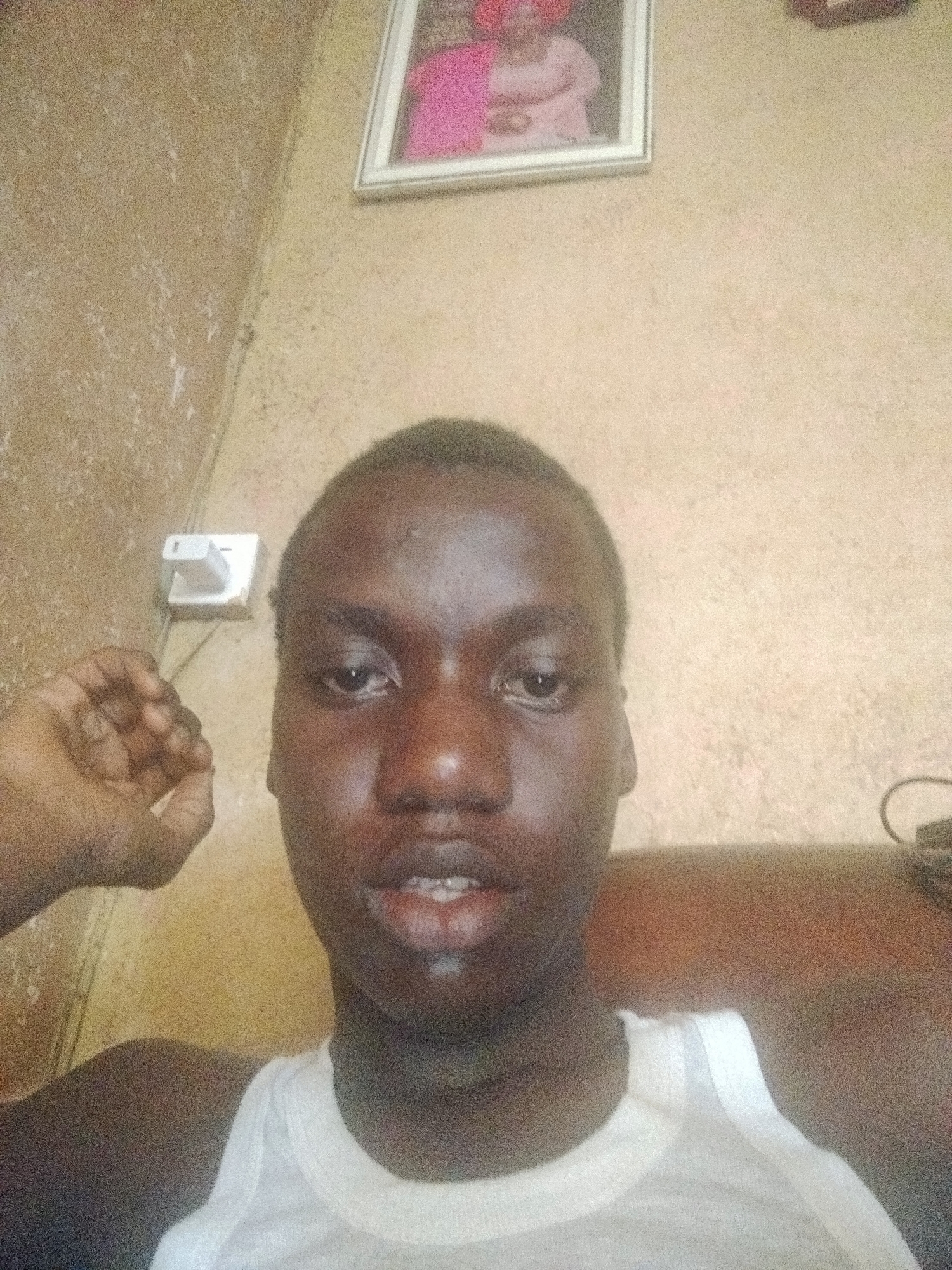 A grainy, eye-level selfie depicts a young individual, likely male, with dark skin and short, closely cropped dark hair. The person is looking directly into the camera with a neutral expression, mouth slightly open. They are wearing a white tank top. Their right hand is partially visible on the left side of the frame, with fingers slightly curled.
The background consists of a textured, light brown or beige wall, which appears to have some wear or marks. On the upper left side of the wall, a white electrical socket with a two-pin plug adapter is clearly visible. In the upper right quadrant of the image, a framed picture hangs on the wall. The picture contains two indistinct figures, one of whom appears to be wearing pink clothing, though details are blurred. A portion of brown furniture, possibly a couch or chair, can be seen behind the person's right shoulder in the lower right corner, with faint hints of cables or wires on it.
The image is dimly lit, contributing to its grainy quality. There is no visible text. The setting appears to be an indoor domestic space, consistent with a home environment in Shangisha, Nigeria.