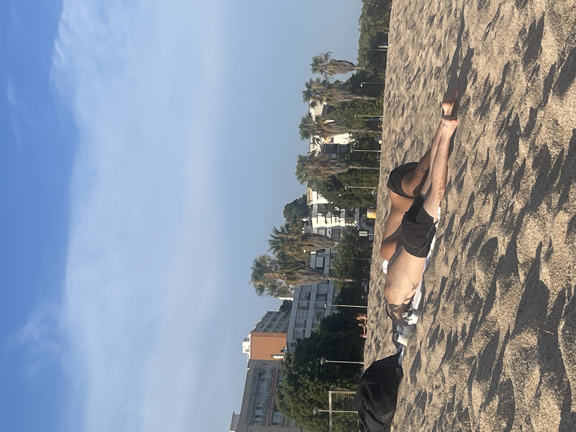 This image depicts a sunny day at the beach in Canet de Mar, Spain. In the foreground, two individuals are lying on the sand, facing away from the camera. One person is wearing black swim shorts and the other appears to be wearing similar attire. A black bag and a pair of white shoes are placed on the sand near them. The beach is sandy with patterns created by the sunlight casting shadows. In the background, there are several buildings, some appearing to be hotels or apartment complexes, with palm trees and other vegetation interspersed. A portion of a soccer goal net is visible on the left side of the image. The sky is blue with scattered white clouds.