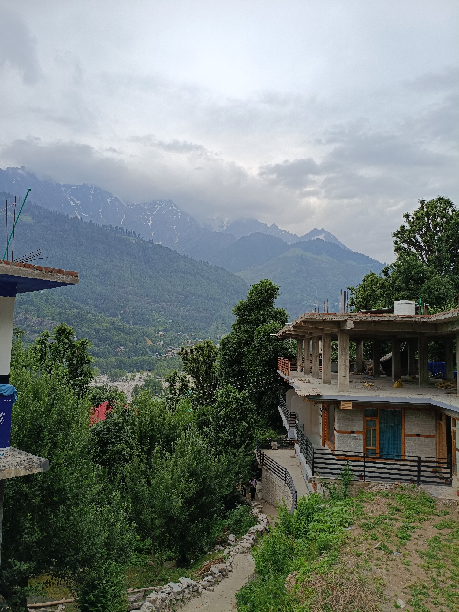 An overcast day in Vashisht, India, presents a view of snow-capped mountains looming behind a lush green valley dotted with buildings. In the foreground, a partially constructed building with concrete pillars and exposed rebar dominates the right side of the frame, overlooking a steep slope covered in vegetation. To the left, another building's roofline is visible, with a blue tarp and some construction materials. A winding path with stone borders leads down from the buildings, and a few figures can be seen walking on it. The overall scene captures a blend of natural beauty and ongoing development in a mountainous region.