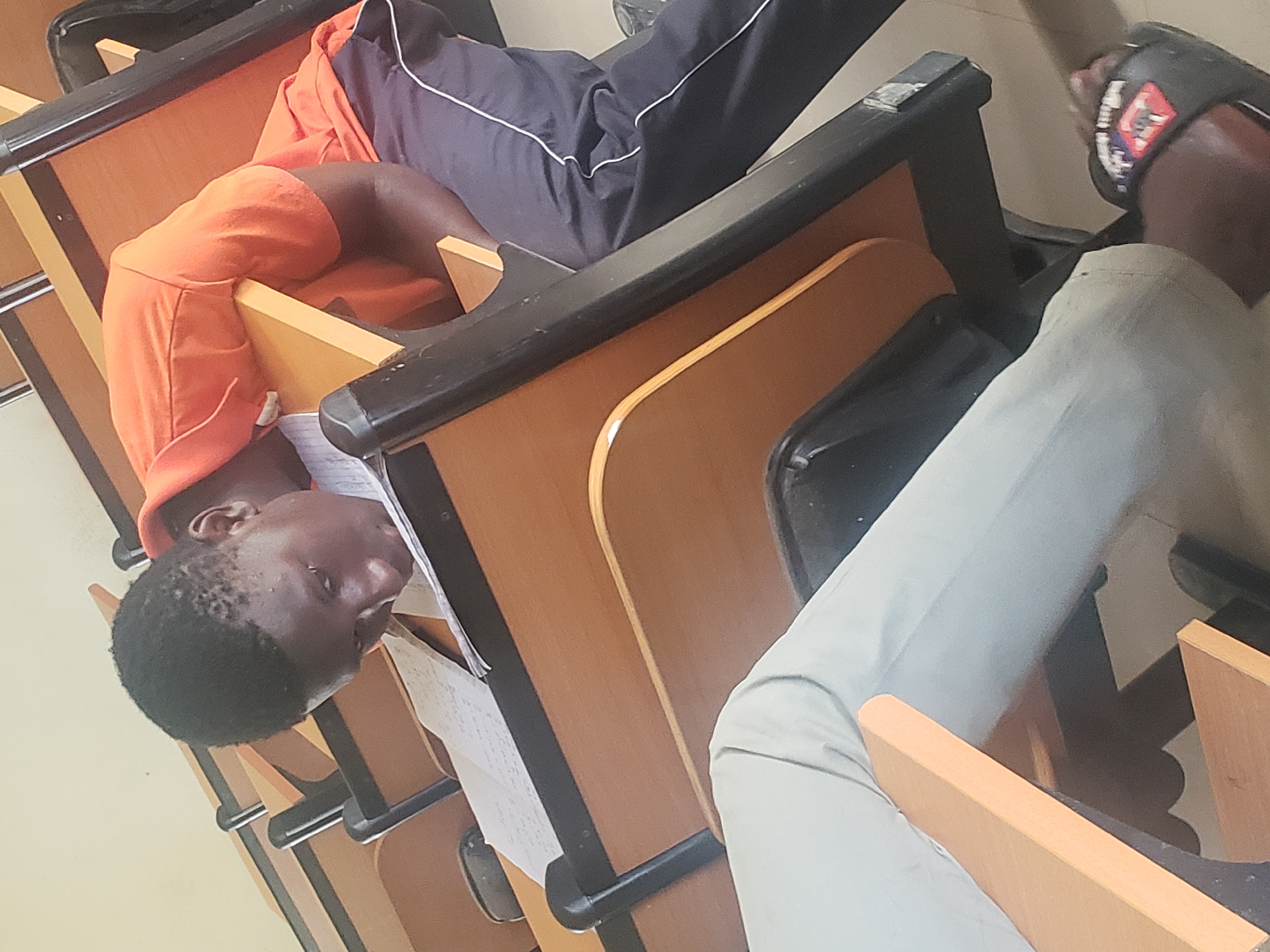 In a classroom in Gumel, Nigeria, a young Black male, likely a student, is seated at a desk, his head lowered as he appears to be sleeping or resting on his open notebook. He is wearing an orange t-shirt and dark sweatpants with white stripes. The classroom features tiered wooden desks with black metal frames. Another student's legs, clad in light-colored pants and black flip-flops, are visible in the foreground, suggesting an occupied classroom environment. The overall scene depicts a typical academic setting, possibly during a lecture or study session.