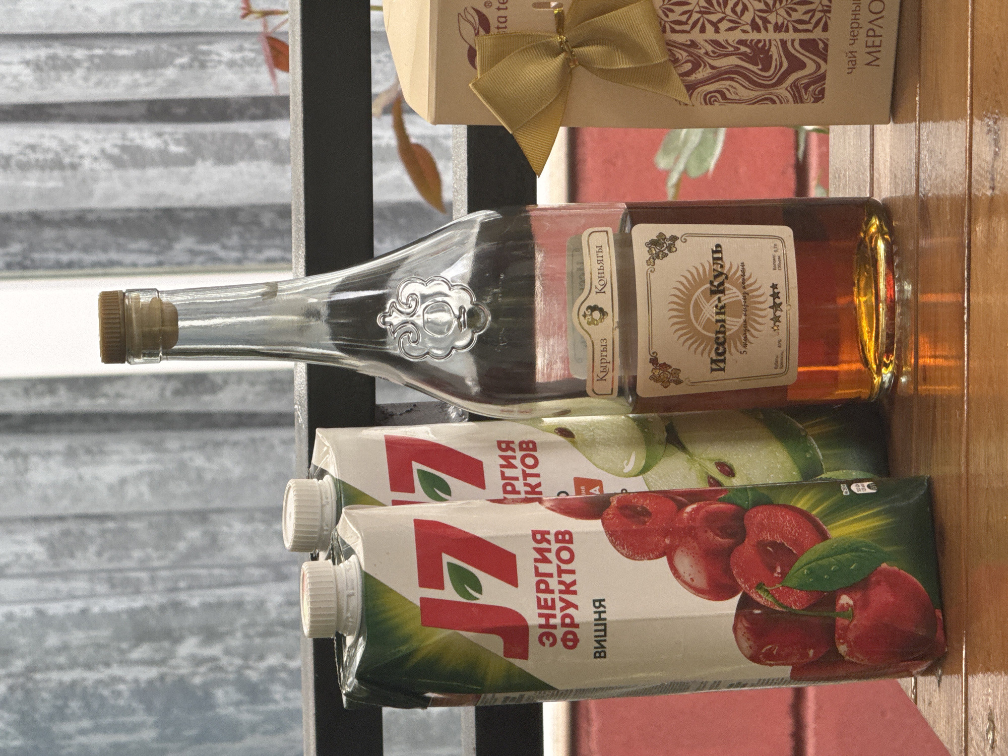 The image depicts a still life arrangement on a wooden surface, likely a table or balcony railing. In the foreground, two cartons of "J7" juice are visible. The carton on the left features an image of cherries and the text "ЭНЕРГИЯ ФРУКТОВ" (Fruit Energy) and "ВИШНЯ" (Cherry). The carton to its right appears to be a different flavor, possibly apple or pear, based on the green fruit depicted.
To the right of the juice cartons stands a clear glass bottle with a cork stopper, containing amber-colored liquid, likely brandy or cognac. The bottle has a decorative silver emblem on its neck and a label that reads "Кыргыз Коньягы" (Kyrgyz Cognac) and "Ыссык-Кул" (Issyk-Kul), indicating its origin. The label also contains star ratings and text in Cyrillic script, including "Крепость. 40%" (Strength. 40%) and "Объем. 0.7л" (Volume. 0.7L).
Further to the right is a small, light-colored gift box with a gold ribbon tied into a bow. The box has a patterned design and some text on its side, which is partially visible and appears to be related to tea, possibly "чай черный" (black tea) and "МЕРЛО" (Merlo).
The background consists of a curtain or blinds, suggesting an indoor or covered outdoor setting. Red and green foliage is visible behind the curtain, and a hint of a red structure is present in the lower left of the frame. The overall lighting suggests it is daytime. The presence of "Issyk-Kul" on the cognac bottle strongly suggests the location is in or near the Issyk-Kul region of Kyrgyzstan, and specifically, given the mention of "Issyk-Kul" as a brand, it is highly likely this image was taken in the city of Sary-Oy, Kyrgyzstan, which is situated on the northern shore of Issyk-Kul lake.