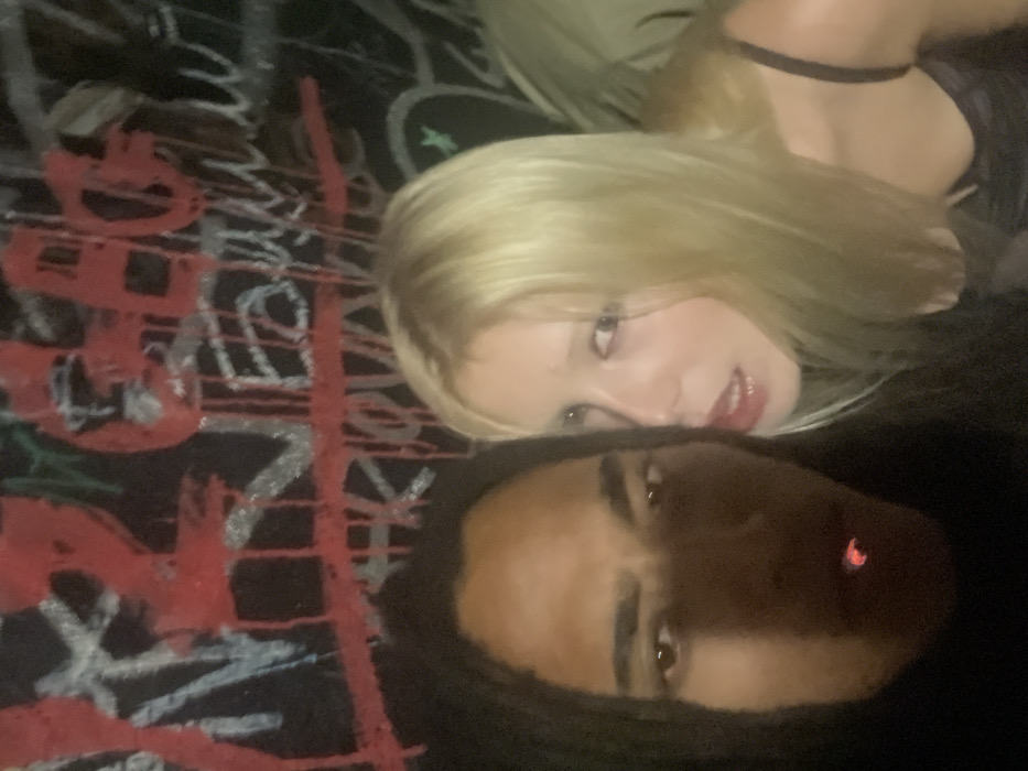 A selfie-style photograph shows two individuals lying down with their faces close to the camera. A blonde woman is positioned above, wearing a black top with a choker, and her gaze is directed towards the camera. Below her, a Black male with dreadlocks is also facing the camera. He appears to be smoking. The background features a surface with graffiti-like artwork in red, white, and other colors. The lighting in the image is somewhat dim.