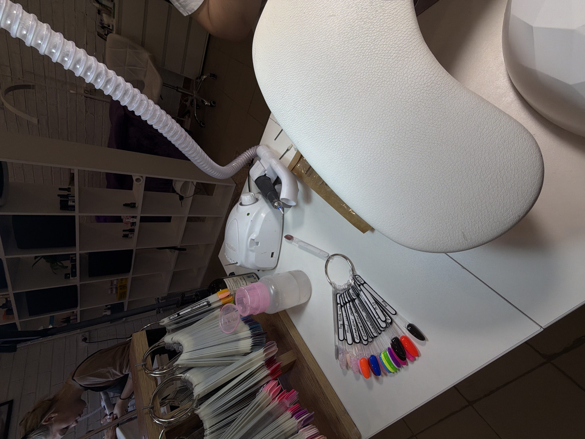 The image shows a manicure station. A white manicure table features a white armrest and a white nail lamp. On the table are a nail drill with a hose, a bottle of nail cleaner, a paintbrush, and a nail color sample ring. A wooden box filled with more nail color samples is also on the table. A person, likely a manicurist, is visible in the lower left corner of the image, and a shelving unit is in the background. The location of this scene is Reshetnikovo, Russia.