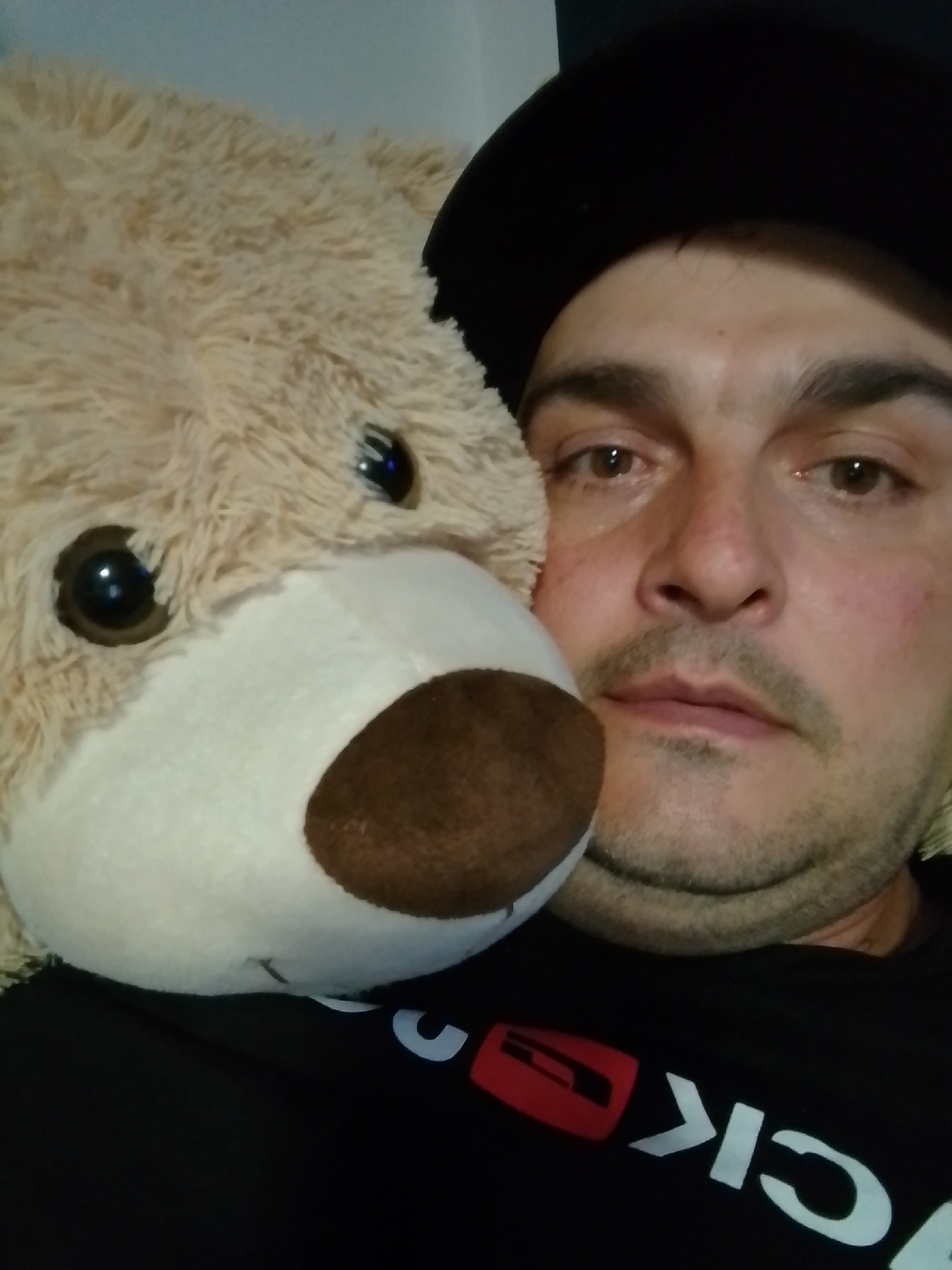 A close-up, overhead shot shows a man and a teddy bear. The man is wearing a black baseball cap and a black t-shirt with white lettering and a red graphic on the front. He has brown eyes, dark hair, and stubble. The teddy bear is light brown and fuzzy, with dark eyes and a brown nose. The man's head is tilted, and he is looking at the camera. The teddy bear is positioned in front of the man, with its head near his face. The background is out of focus, but appears to be indoors. The image was taken in Wedel, Germany.