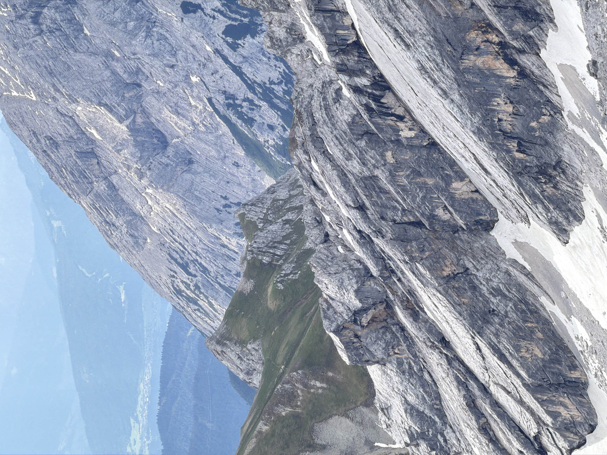 This image captures a layered, mountainous landscape near Garmisch-Partenkirchen, Germany, likely a view from a higher elevation looking towards the Zugspitze massif. The foreground is dominated by jagged, grey rock formations with patches of snow and scree, indicating a high-altitude, rugged terrain. These formations are angled steeply, suggesting a mountain slope.
Further back, rolling green hills transition into darker, forested slopes, and in the distance, a hazy valley with a small settlement is visible. The furthest peaks are bathed in a soft blue light, fading into the sky. The overall impression is one of vastness and natural grandeur, typical of the Bavarian Alps. No people or specific objects are discernible in the image.