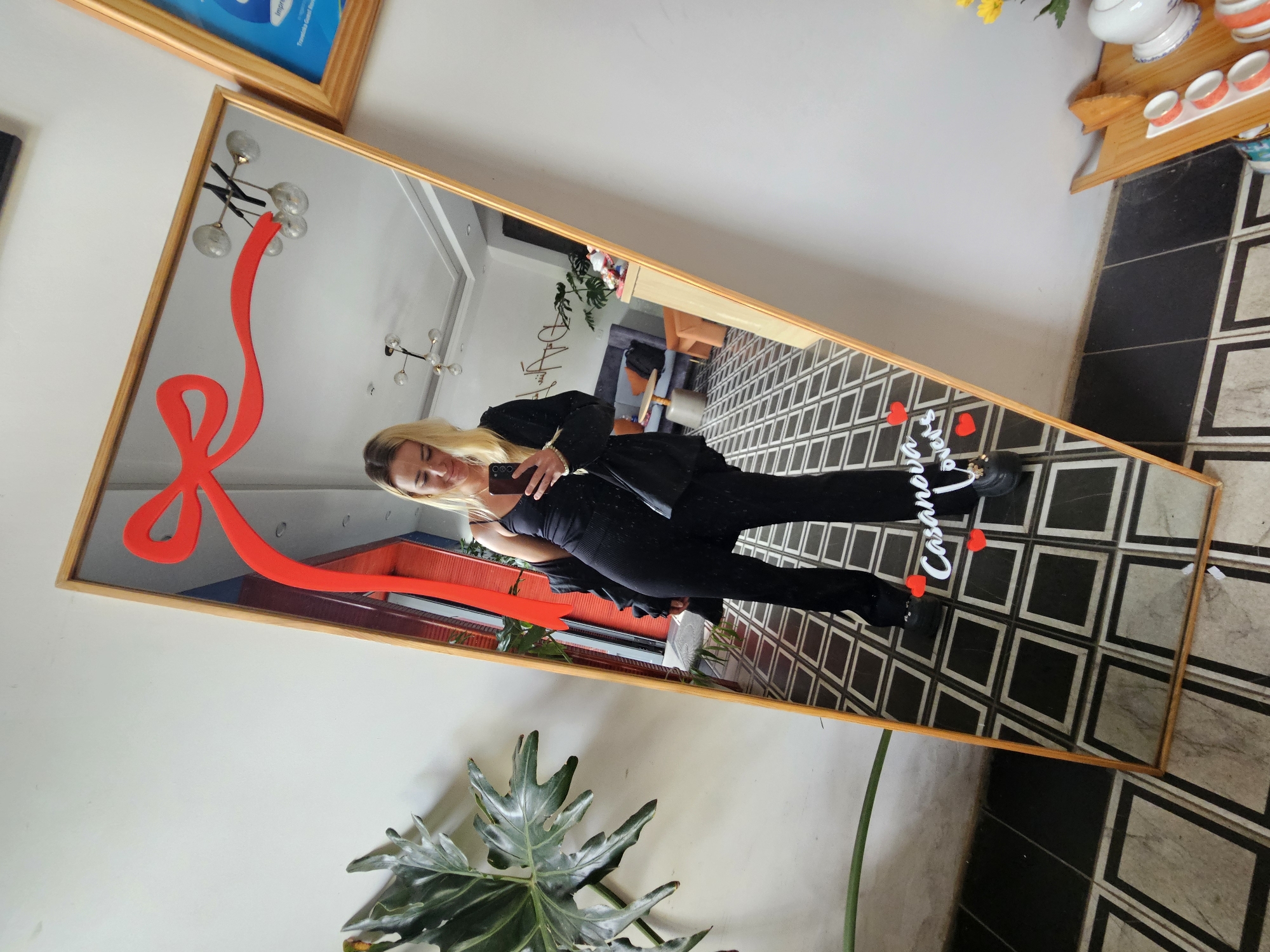 In Da Lat, Vietnam, a full-length mirror with a gold frame stands against a white wall. A red ribbon graphic is adhered to the top of the mirror, with a bow and trailing ends. A woman with blonde hair, wearing a black fitted tank top, black flared pants, and a black jacket draped over her shoulders, takes a mirror selfie with her smartphone. She is wearing black shoes. Two small red heart decals are placed on the mirror near her feet. The floor in front of the mirror has a black and white tiled pattern. Reflections in the mirror show a brightly lit interior with modern furniture, including sofas and a coffee table, a large plant, and a unique chandelier on the ceiling. A framed blue and white poster hangs on the wall to the right of the mirror, and a wooden shelf with small white cups and a teapot is visible on the right side of the image. A large, leafy green plant is positioned to the left of the mirror.