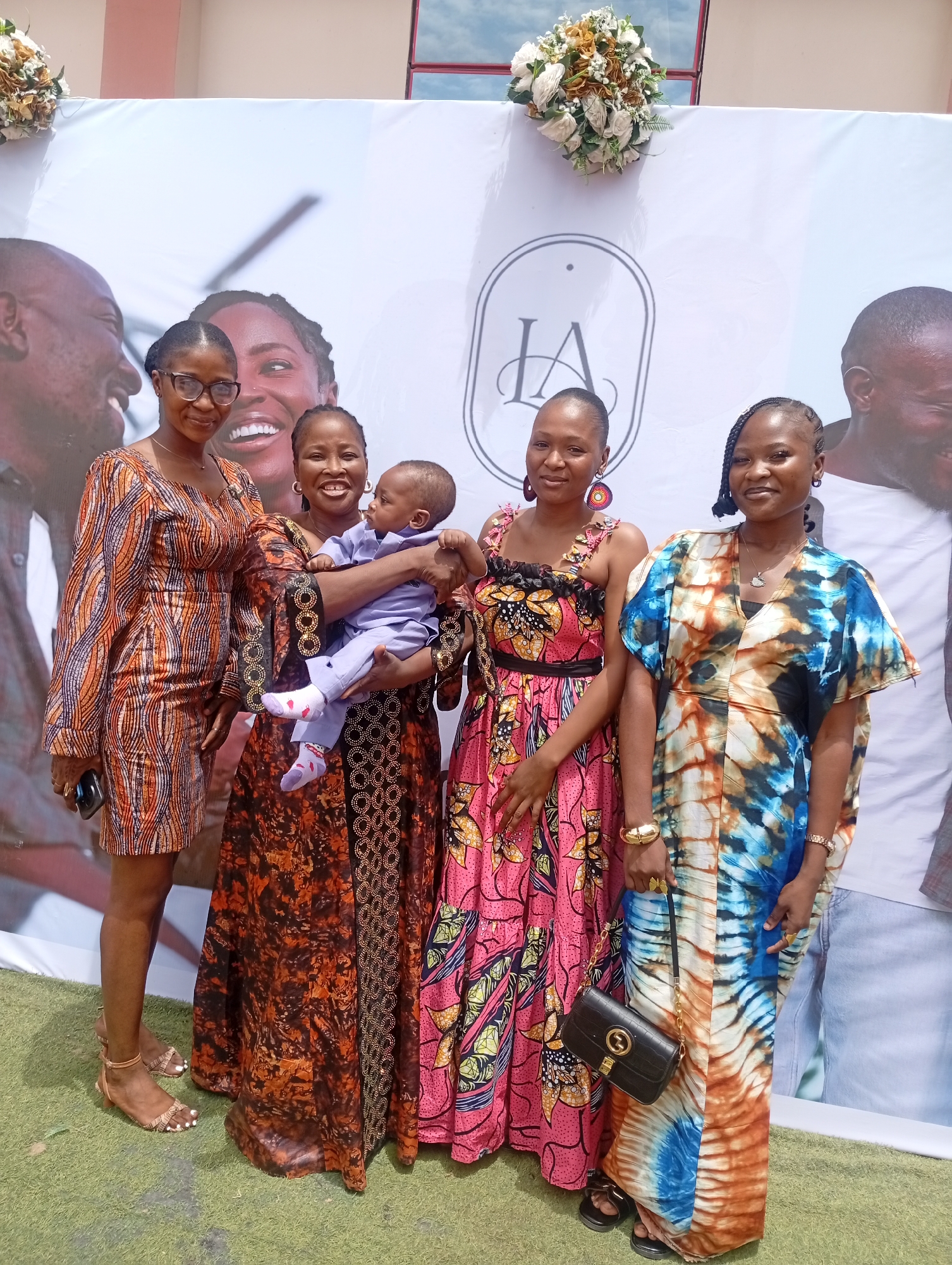 This image, captured in Sabon Tasha, Nigeria, depicts four adult females and one infant standing side-by-side in front of a large white banner. The banner features faded grayscale images of smiling adults, including a man in profile on the left, a woman's face in the center, and a man in profile on the right. A white oval logo with the stylized letters "IA" is positioned in the upper center of the banner. Two floral arrangements, composed of white, cream, and light brown flowers with green foliage, are attached to the top corners of the banner. Above the banner, the light pink-beige wall of a building and a red-framed window are visible. The ground in the foreground is covered with artificial green grass. From left to right: 1. **First Woman:** An adult female with dark skin, wearing dark-rimmed glasses and a short-sleeved, knee-length dress patterned with orange, brown, and blue abstract lines. She holds a dark-colored smartphone in her left hand. Her feet are visible, adorned with light brown multi-strap sandals and white-painted toenails. She is smiling towards the camera. 2. **Second Woman:** An adult female with dark skin, wearing gold-colored hoop earrings and a long, dark brown dress featuring a distinct pattern of orange and black circular motifs on the lower half. She is smiling towards the camera while holding an infant in her arms. 3. **Infant:** A baby, appearing to be approximately 6-12 months old, dressed in a light blue two-piece outfit with white socks patterned with pink accents. The infant is facing towards the right side of the frame, not looking directly at the camera. 4. **Third Woman:** An adult female with dark skin, wearing large, circular drop earrings with red, blue, and yellow accents. She wears a sleeveless, long dress with a predominantly pink, orange, and black floral and abstract pattern. The bodice has a ruffled design. She has a gold-colored bracelet on her left wrist and is smiling towards the camera. 5. **Fourth Woman:** An adult female with dark skin and braided hair, wearing a loose-fitting, long kaftan-style dress with a blue, orange, brown, and white tie-dye pattern. She carries a black handbag with a gold chain strap and a gold-toned "G" logo in her left hand. She wears a gold-colored watch on her left wrist, a gold-colored bangle on her right wrist, and a gold-colored ring on her right hand. Her feet are partially visible
