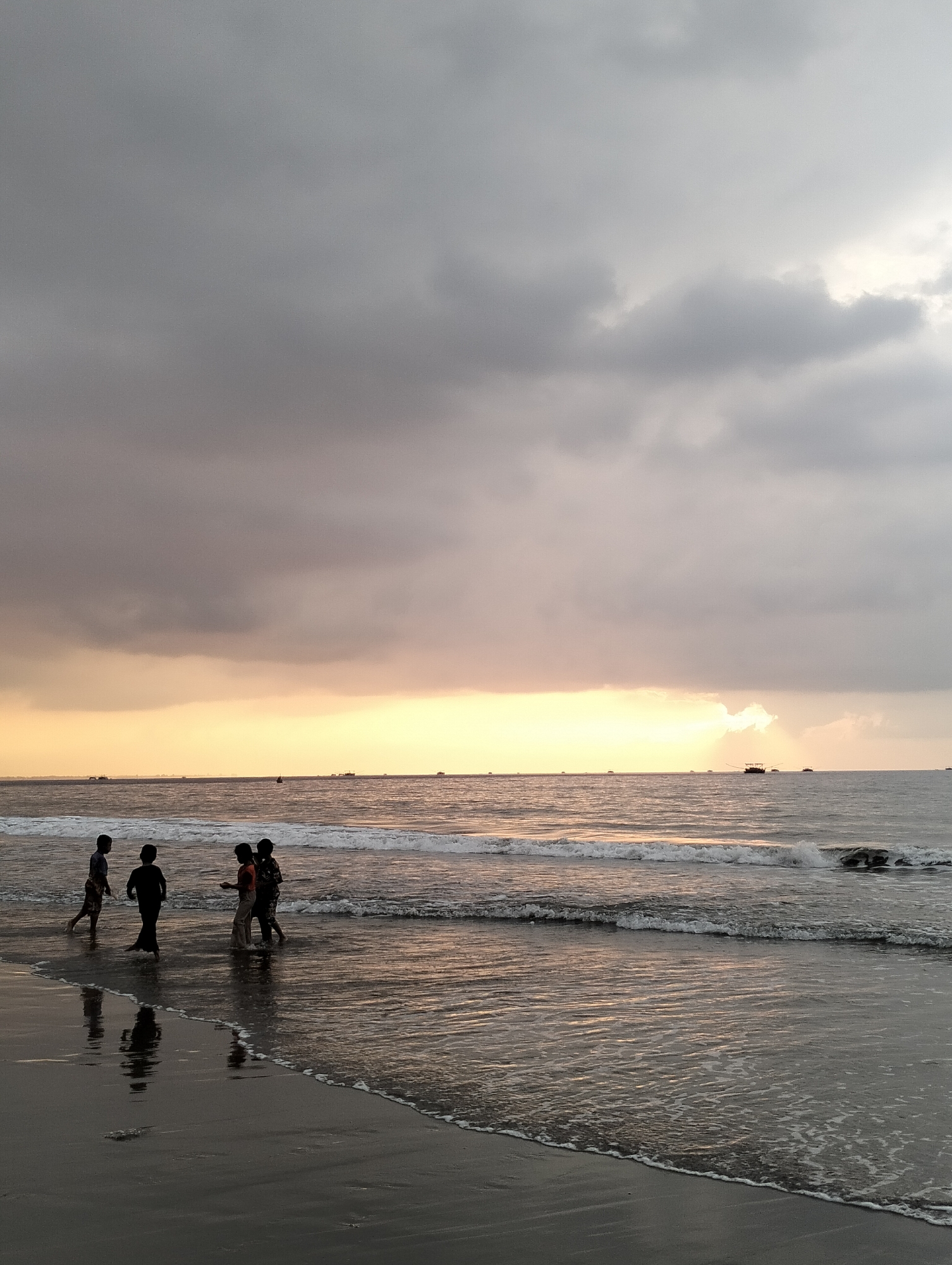 The image captures a serene beach scene in Peuneulet Baroh, Indonesia, during a cloudy sunset. Four individuals, silhouetted against the dusky sky, stand in the shallow water near the shore. Two of them appear to be children, while the other two are possibly older. They are positioned close to each other, engaged in what seems like a casual interaction. Gentle waves lap the dark sand, creating a reflective surface that mirrors the figures and the overcast sky. In the distance, several boats are visible on the calm sea, adding a touch of activity to the otherwise tranquil setting. The horizon is marked by a soft gradient of color, transitioning from a pale yellow glow where the sun is setting, to the grays and blues of the impending evening. The overall atmosphere is one of peaceful solitude and natural beauty.