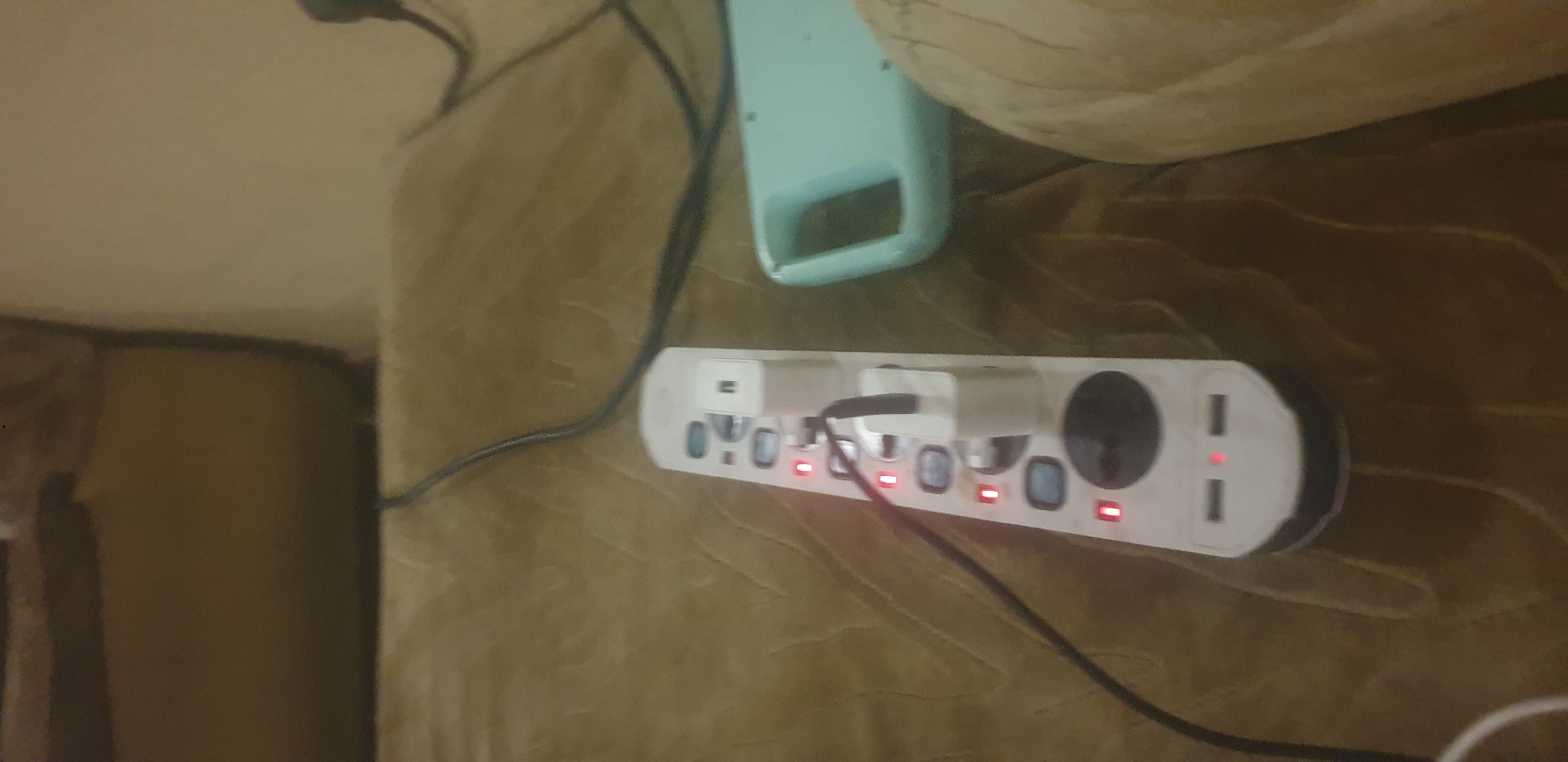 The image shows a close-up of a white power strip with multiple outlets and USB ports, situated on a brown textured surface, likely a sofa or carpet. Several red indicator lights are illuminated on the power strip, suggesting that devices are plugged in and receiving power. Two white power adapters are plugged into the power strip, one of which has a black cable attached. To the right of the power strip is a light blue object with a handle, possibly a storage container or electronic device. The background is blurred, but appears to be indoors. There are no people visible in the image. The location is Unknown, Unknown.