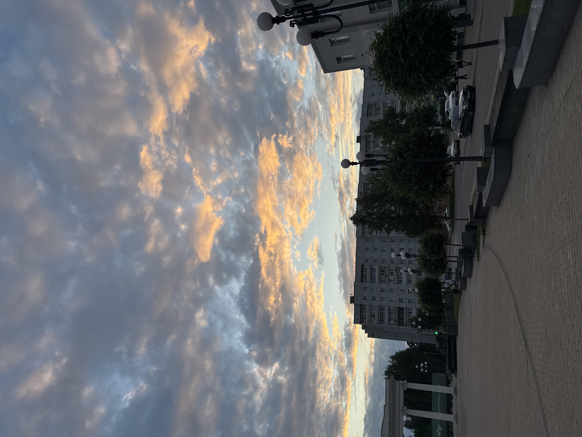 The image shows a street scene in Chaikovsky, Russia, with a large residential building in the background. The sky is filled with dramatic clouds illuminated by the warm colors of sunset, ranging from soft oranges and yellows to cooler blues and grays. In the foreground, a paved walkway curves to the right, bordered by concrete benches and trees. A street with several cars is visible, including a dark sedan in the middle of the road. Streetlights with multiple globes are positioned along the walkway. To the left, a white arched structure with columns stands near some trees and a green train car. People are walking on the sidewalk in the distance. The overall atmosphere is one of a city transitioning into evening under a vibrant sky.