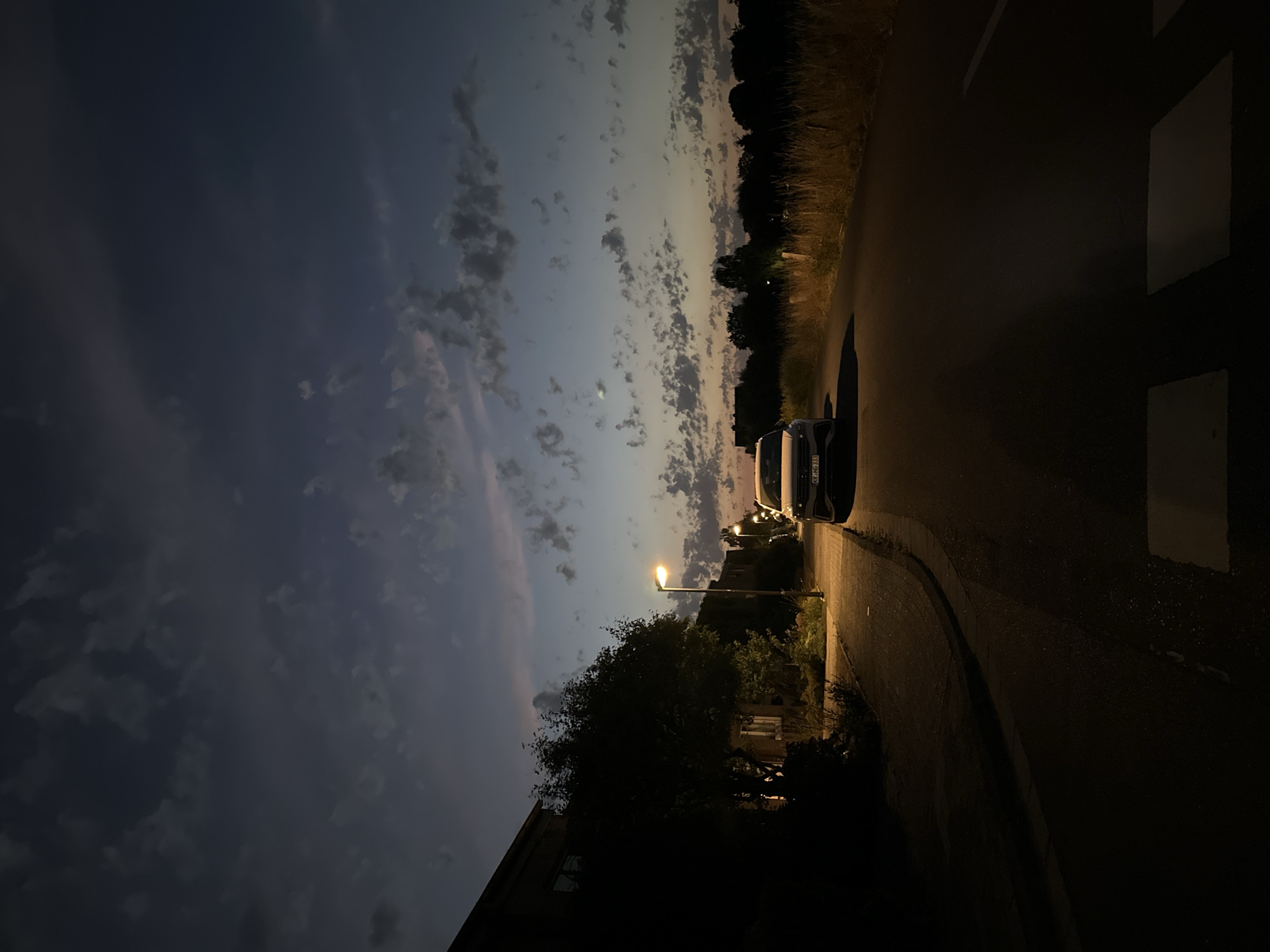 The image depicts a street scene in Krefeld, Germany, during dusk. A white car is parked on the right side of the asphalt road, facing away from the viewer. The car's license plate reads "KR-NG 333". To the left of the road, there is a sidewalk with a lamppost illuminating the area. Further left, a dark building with windows and a tree are visible. The sky is a deep blue with scattered clouds, some tinged with pink from the setting sun. The environment appears to be residential, with houses lining the street. The overall mood is calm and serene, typical of a quiet evening in a suburban area.