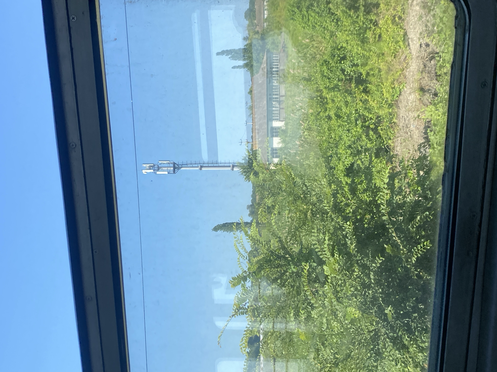 The image depicts a view through a train window, showcasing a rural landscape under a clear blue sky. In the foreground, lush green foliage, likely bushes and trees, partially obscures the view of a low-slung building with a grey roof, suggesting it might be a single-story structure. Behind the building, a tall telecommunications tower with multiple antennas stands prominently, its white structure contrasting with the sky. The overall scene suggests a typical daytime view from a moving train, passing through a sparsely populated area that appears to be in or near the city of Azadogly, Russia. The lighting indicates it is daytime, likely with strong sunlight given the clear sky. There are no people visible in the frame.