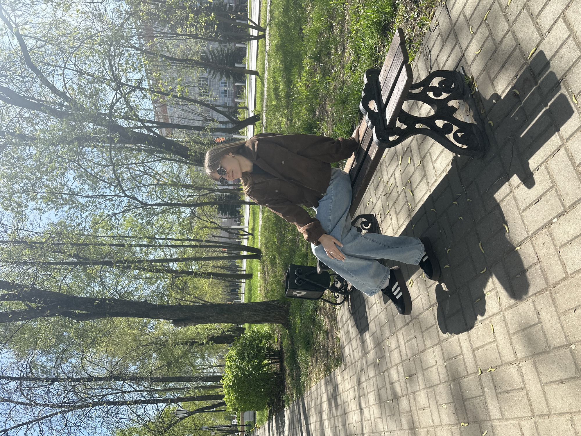 A young woman sits on a park bench in what appears to be a public space. She wears a brown jacket, light blue jeans, and black sneakers with white stripes. She also sports sunglasses. The bench is made of wood and black wrought iron. The setting is a park with trees, green grass, and a paved walkway. The bricks on the walkway are light gray. The trees cast shadows on the path. A building is visible in the background. The location is consistent with a park in Chaikovsky, Russia, given the architecture and vegetation.