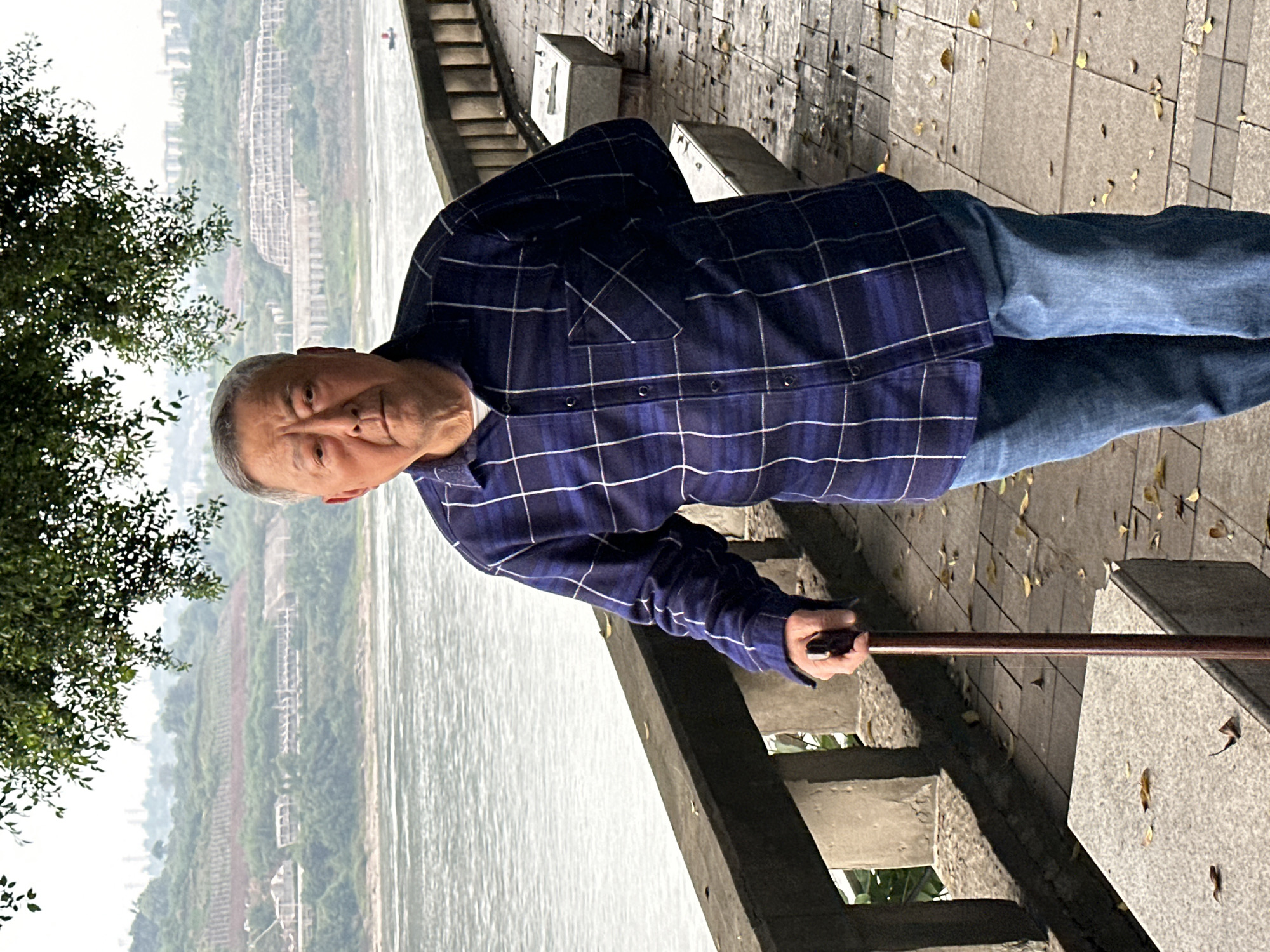 An elderly Asian man stands on a concrete pathway along a riverbank in Jiangjin, China. He is wearing a blue and white plaid shirt and blue jeans, holding a brown cane. The pathway is made of large concrete tiles scattered with fallen yellow leaves and lined with a low concrete barrier. Across the river, a hazy cityscape is visible, with buildings lining the hills. A tree with green foliage is present on the left side of the image.