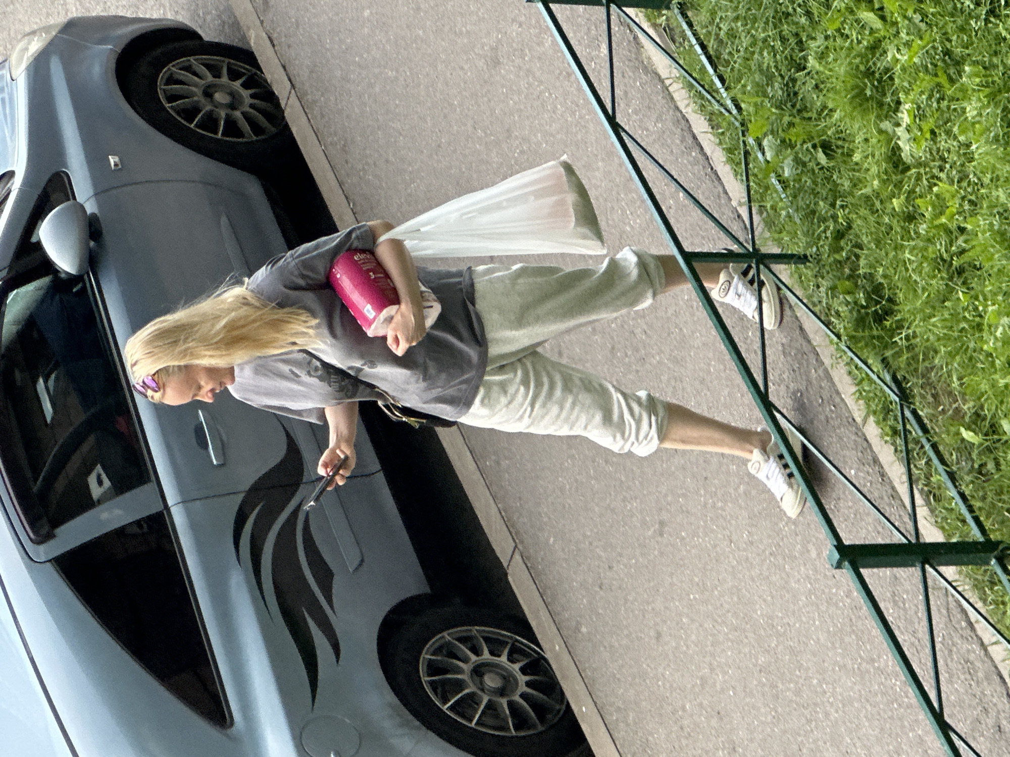 In Murino, Russia, a woman with blonde hair, wearing a grey t-shirt, light-colored sweatpants, and white athletic shoes, walks on a paved sidewalk. She is holding a pink cylindrical object, likely a roll of toilet paper, in her left hand and a white plastic bag with some items inside in her right hand. Her gaze is directed downwards at a mobile phone she holds in her right hand. To her left is a light blue car parked alongside the sidewalk, featuring a black flame decal on its side. The scene is bordered by a green metal fence in the foreground and lush green grass to the right.