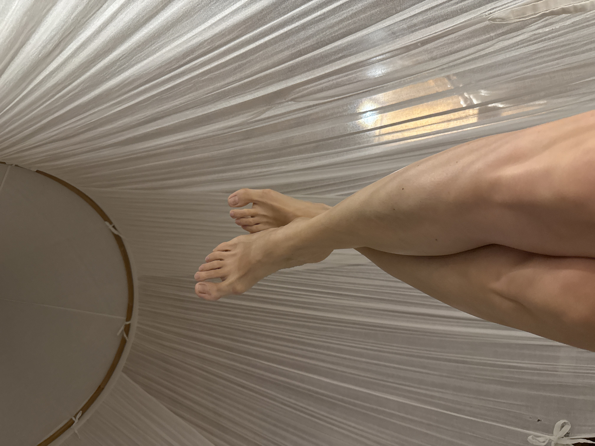 The image presents a partial view from within a white fabric structure, possibly a mosquito net or canopy. In the foreground, a pair of crossed legs and bare feet are visible. The fabric is stretched and pleated, obscuring the background but suggestive of an interior space, possibly a bed. The lighting is soft and diffused. The location, Mengwi, Indonesia, cannot be confirmed from the visual information provided.