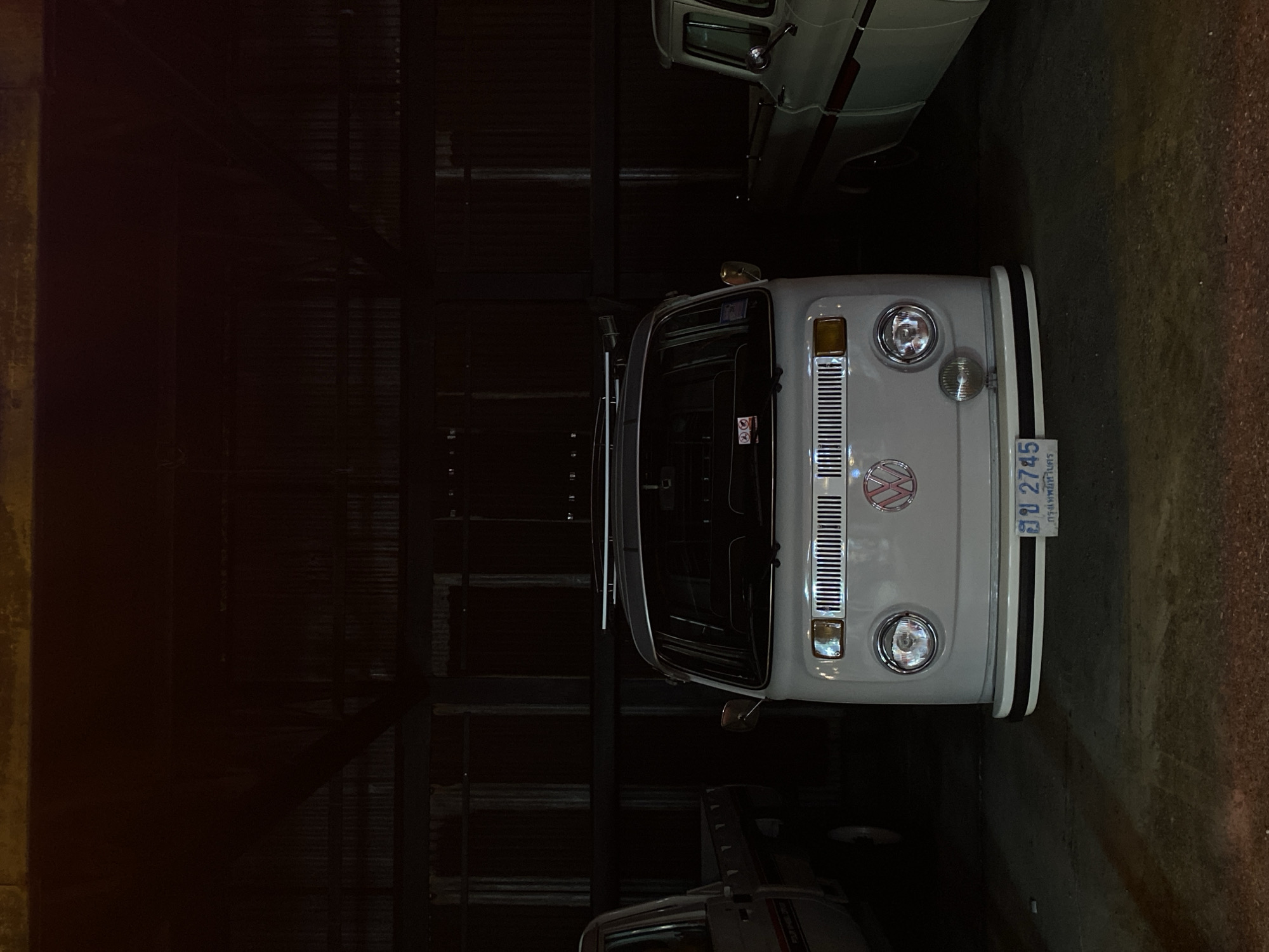 The image features the front of a vintage Volkswagen van, recognizable by its distinctive round headlights and VW emblem. The van is light gray in color. The license plate reads "อย 2745" and appears to be a Thai license plate, indicating that the location is in Thailand. The writing on the license plate includes the word "กรุงเทพมหานคร", which is the Thai name for the city of Bangkok. The van seems to be parked indoors, possibly in a garage or storage facility, as suggested by the dark and enclosed environment with metal bars or structures in the background. Other vehicles are also partially visible behind and beside the van. The light in the scene is dim.