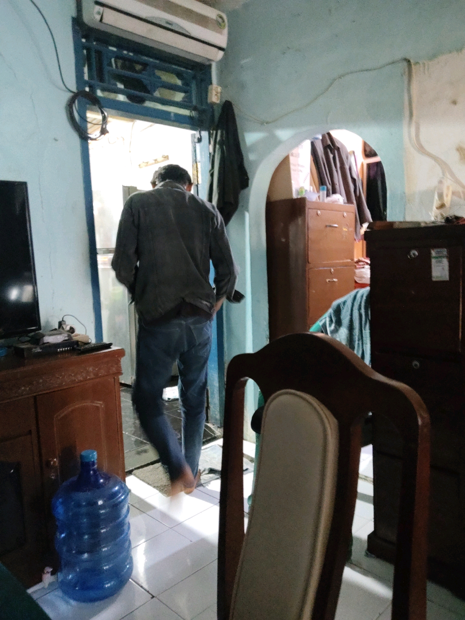 A man in a dark jacket and blue jeans walks away from the camera towards an open doorway in a room with light blue walls. The doorway leads to a brightly lit outdoor area. To the left of the doorway, a flat-screen television is visible on top of a wooden cabinet. In the foreground, a large blue water jug with a white spigot sits on the floor. A wooden chair with a light-colored cushion is in the right foreground, partially obscuring a dark wooden cabinet. In the background, through an arched opening, several large brown filing cabinets are stacked. The scene appears to be an interior residential or small office space in Subang, Indonesia.